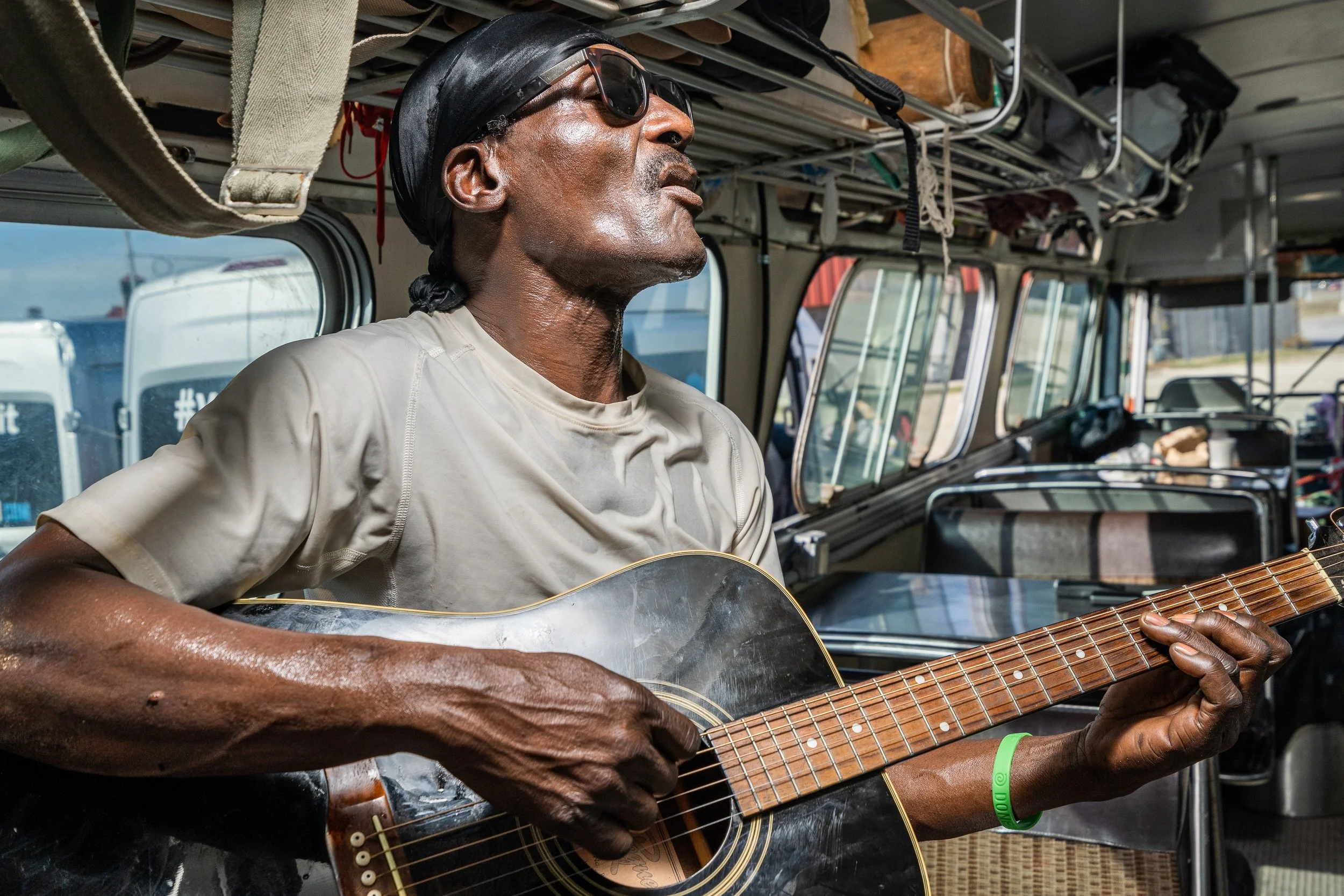 A man wearing sunglasses and a black head covering is playing an acoustic guitar inside a vehicle, possibly a bus, with overhead storage and windows showing parked vehicles outside.