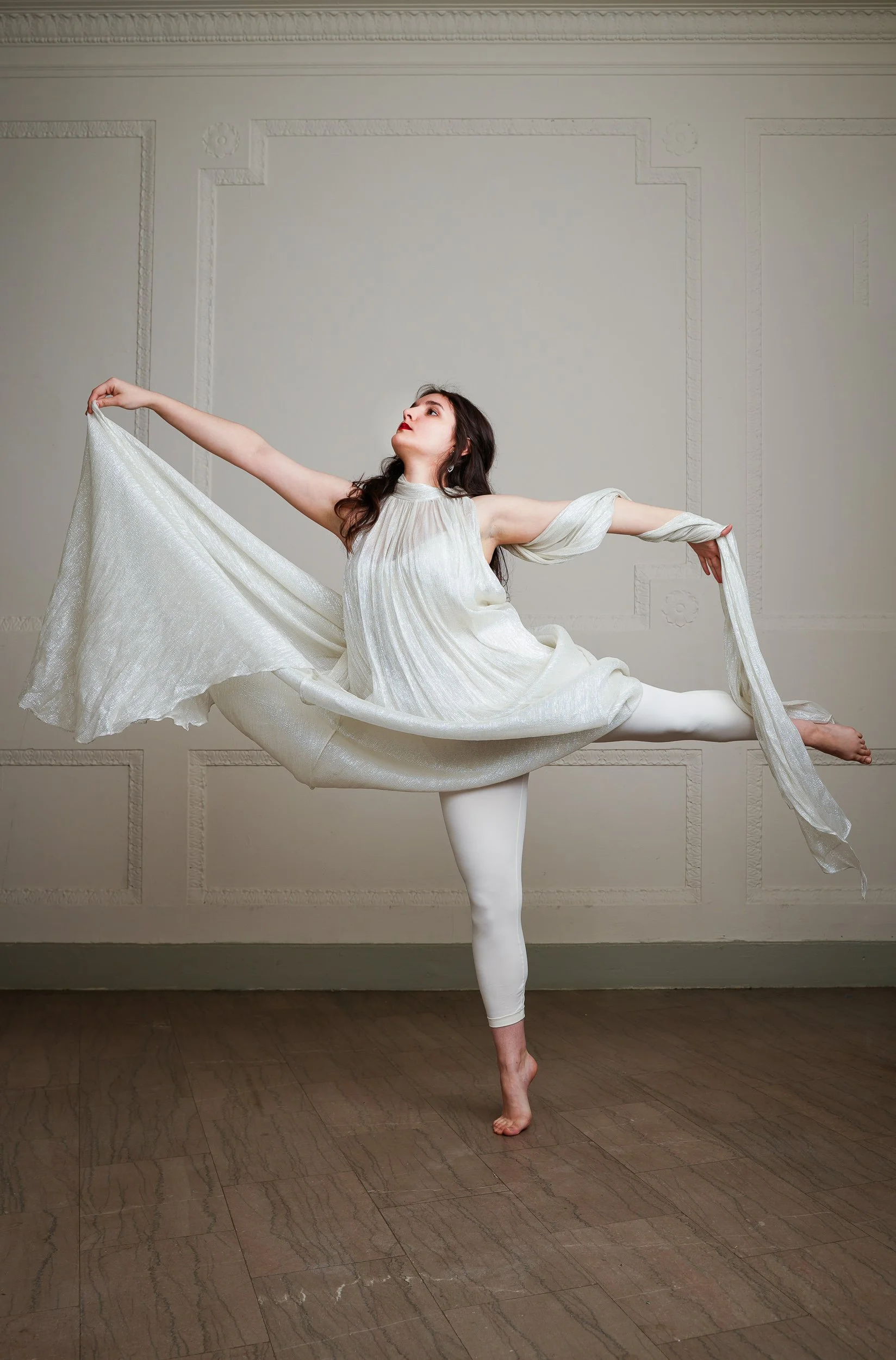 A woman performing a ballet pose on one leg in a room with decorative wall panels, wearing white tights and a flowing white dress with draped sleeves.