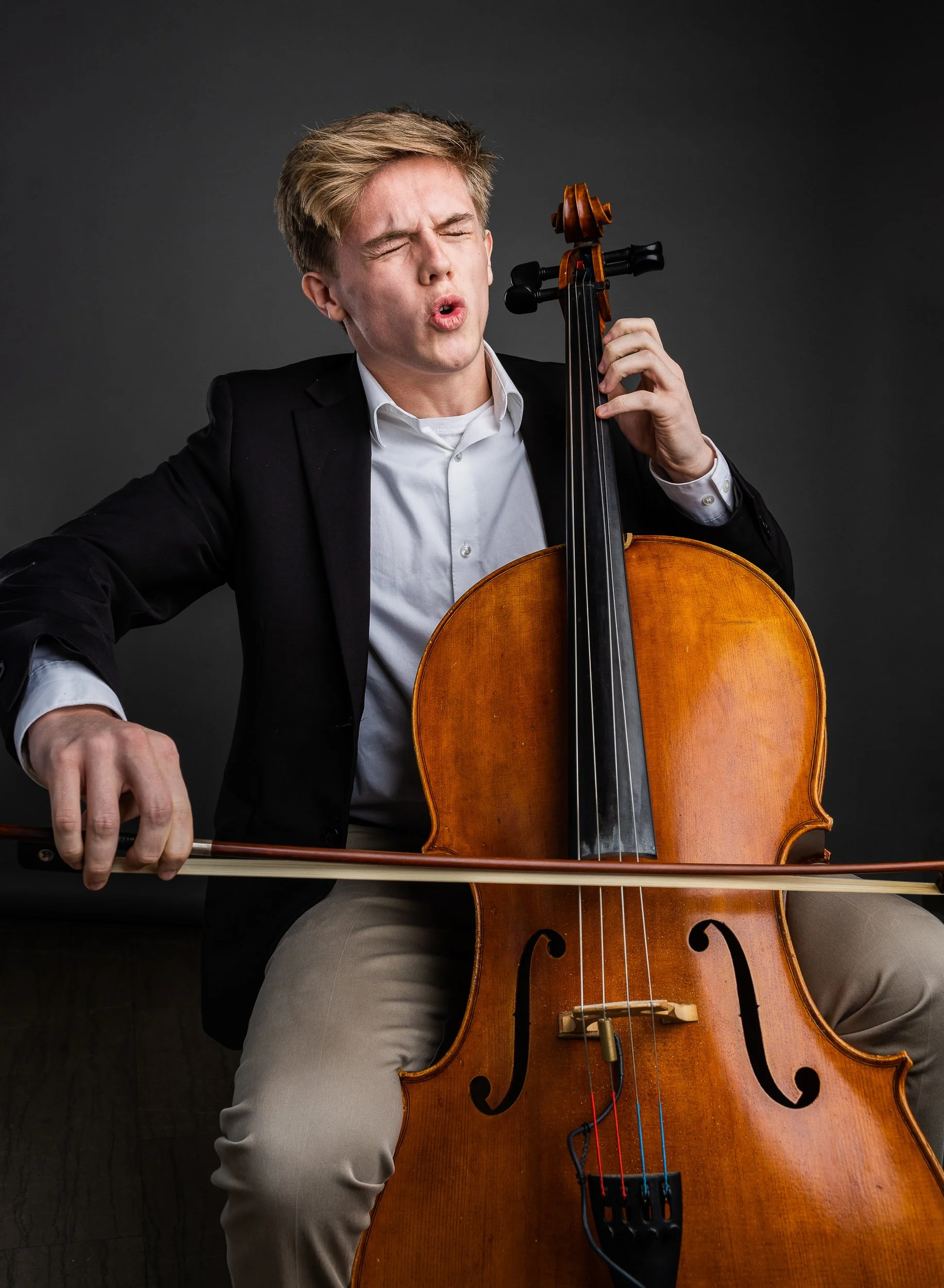 A young man with blonde hair wearing a black blazer and white shirt playing a cello with a bow, eyes closed, expressing emotional engagement, set against a dark background.