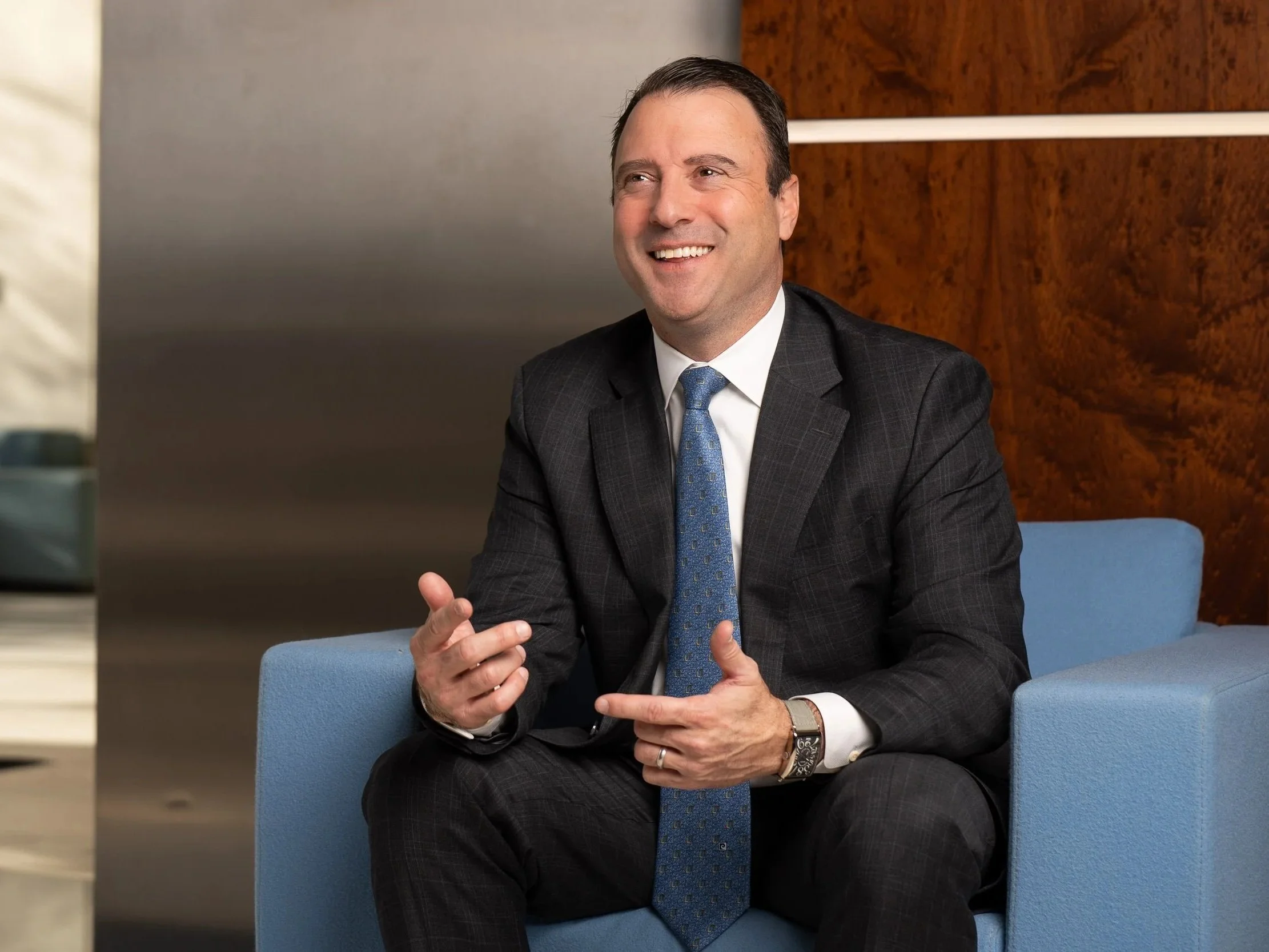 A man in a dark gray suit with a light blue tie, smiling, sitting on a blue chair in a modern office setting with wood and gray walls.