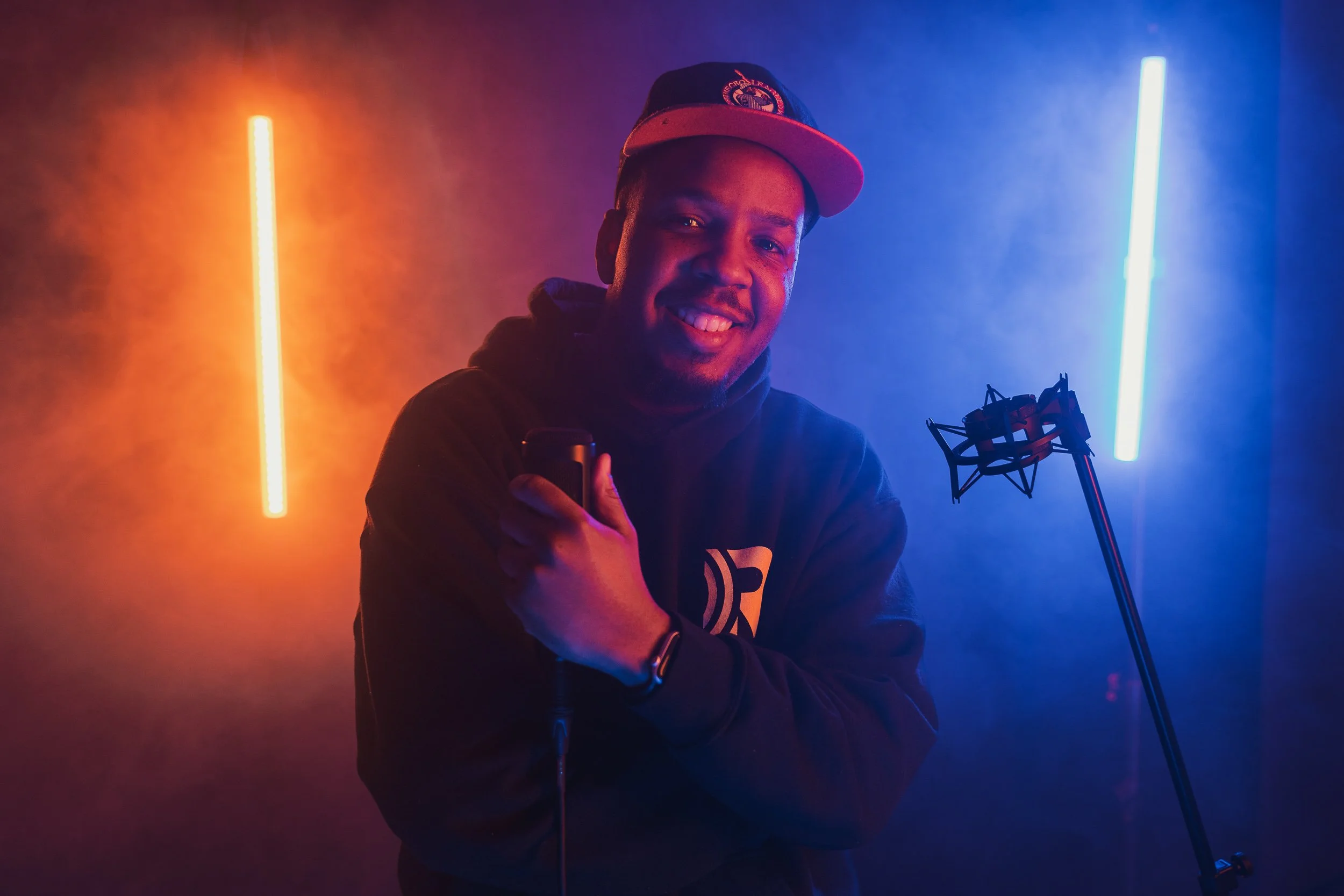 A young man smiling, wearing a black hoodie and a baseball cap, standing in front of colorful neon lights with blue and orange hues. He is holding a smartphone in his right hand and there is a studio microphone on a stand beside him.