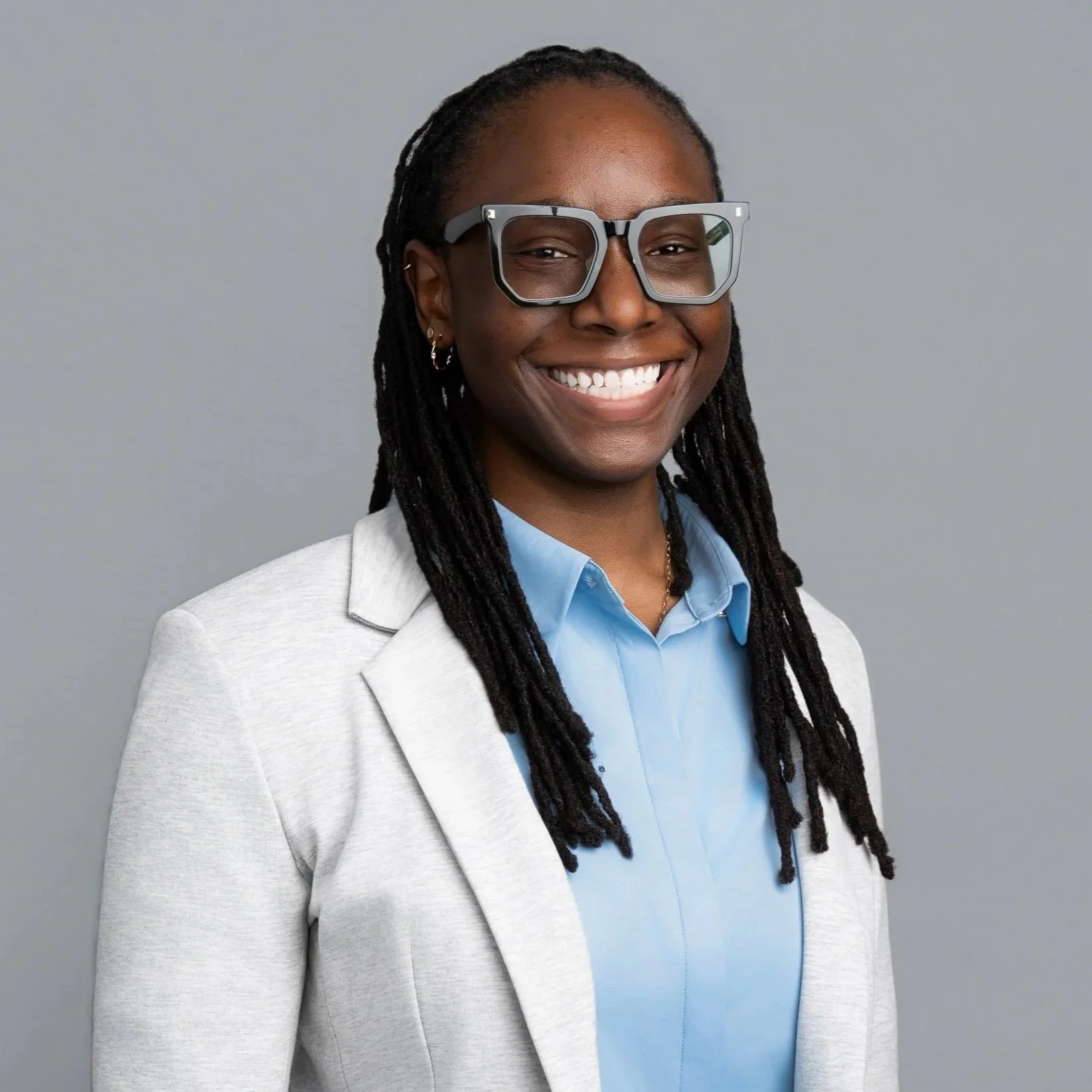Portrait of a smiling woman with long dreadlocks, wearing large gray glasses, a light gray blazer, and a blue collared shirt, standing against a gray background.
