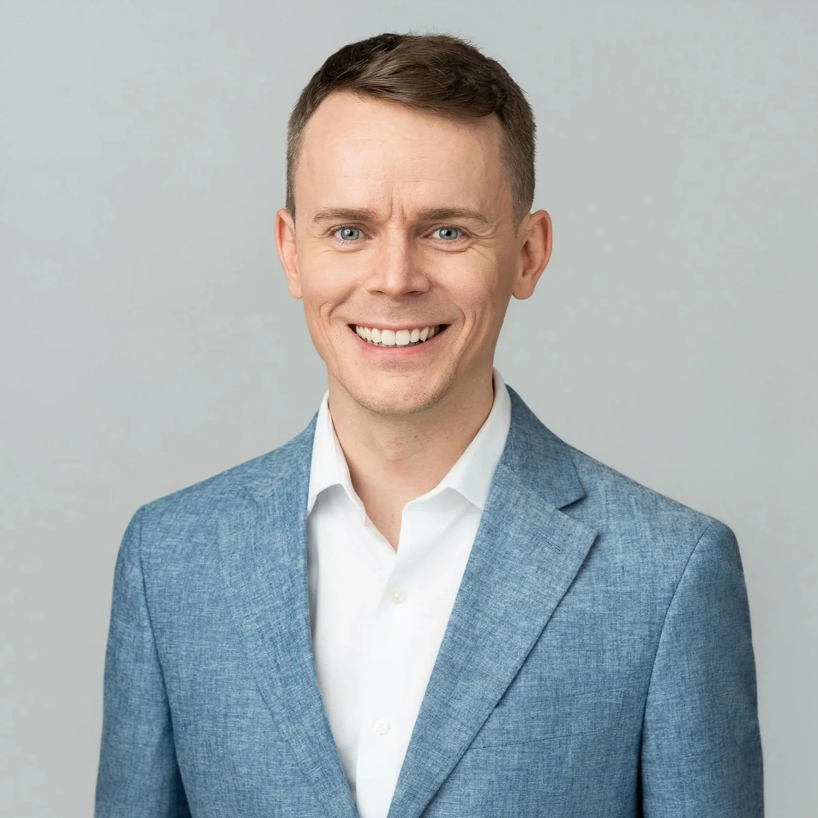 A young man with short brown hair, blue eyes, and a wide smile, wearing a blue blazer over a white shirt, posed against a plain gray background.