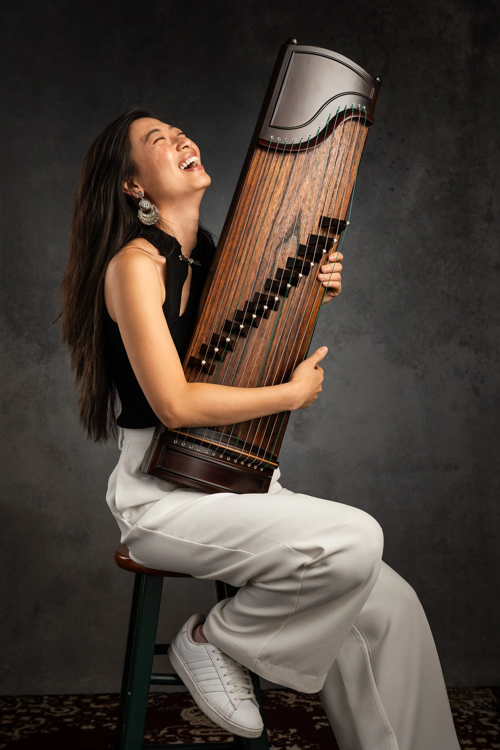Woman dressed in black top and white pants sitting on a stool, playing a large hammered dulcimer, laughing joyfully against a dark textured background.