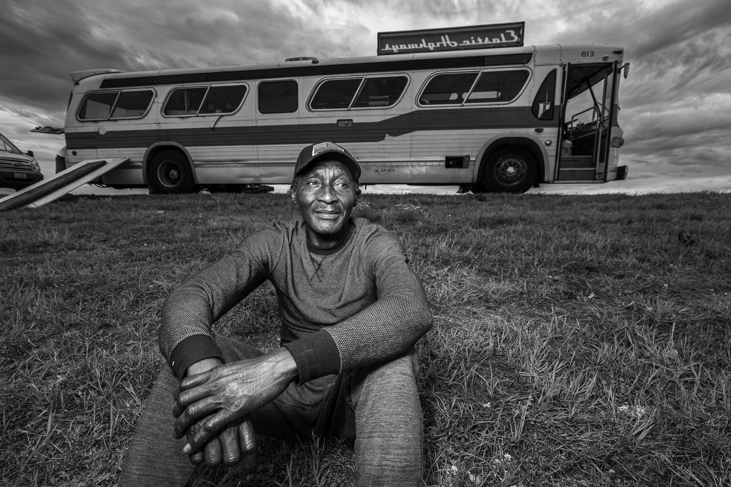 A man sitting on the grass with a bus and parked cars behind him under cloudy skies.