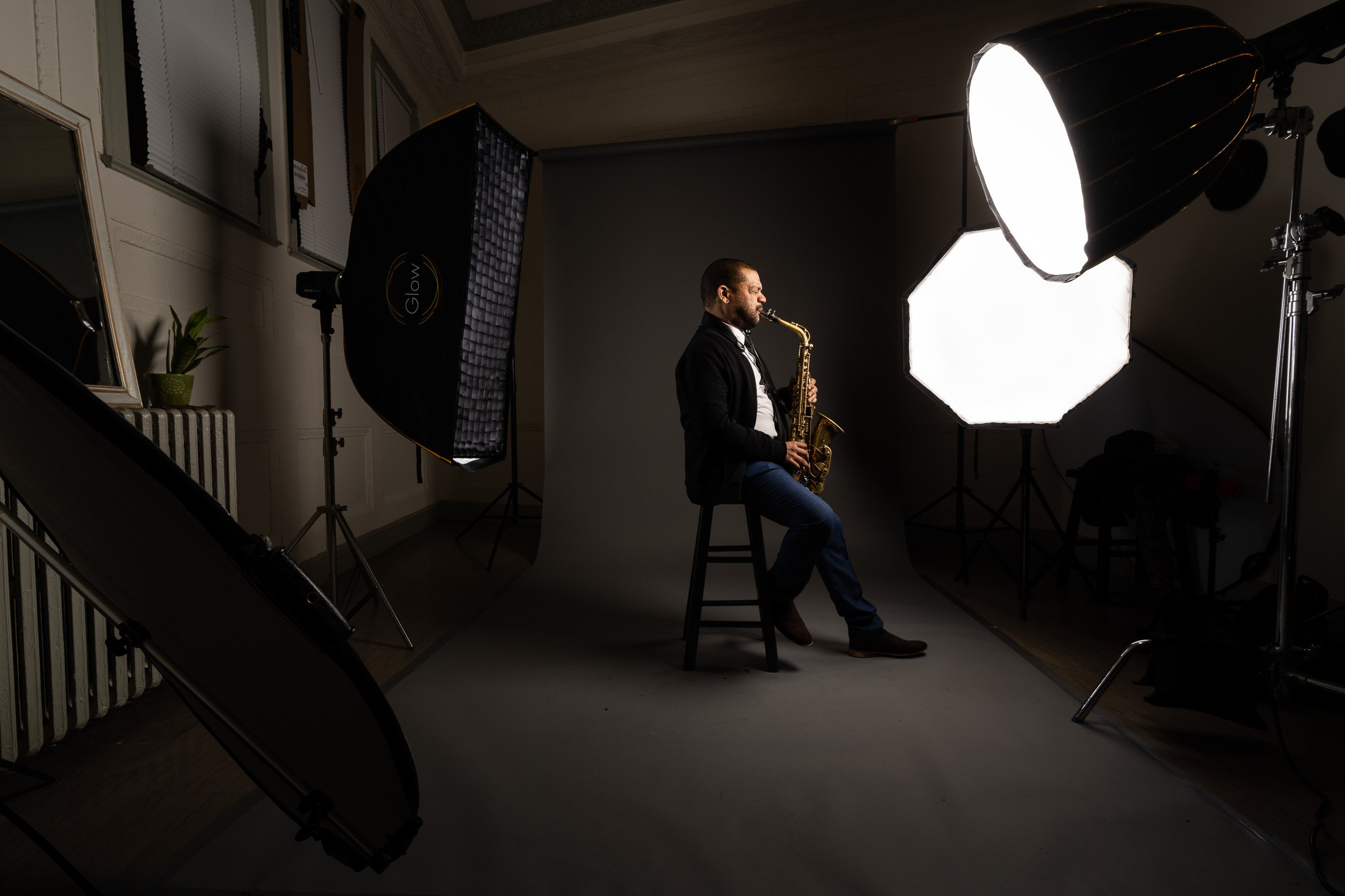 A man in a black jacket and blue jeans is sitting on a stool in a photography studio playing a saxophone. The studio is equipped with large professional lights, including a white softbox and a large reflector, directed at the man. There are mirrors, a plant, and window blinds visible in the background.