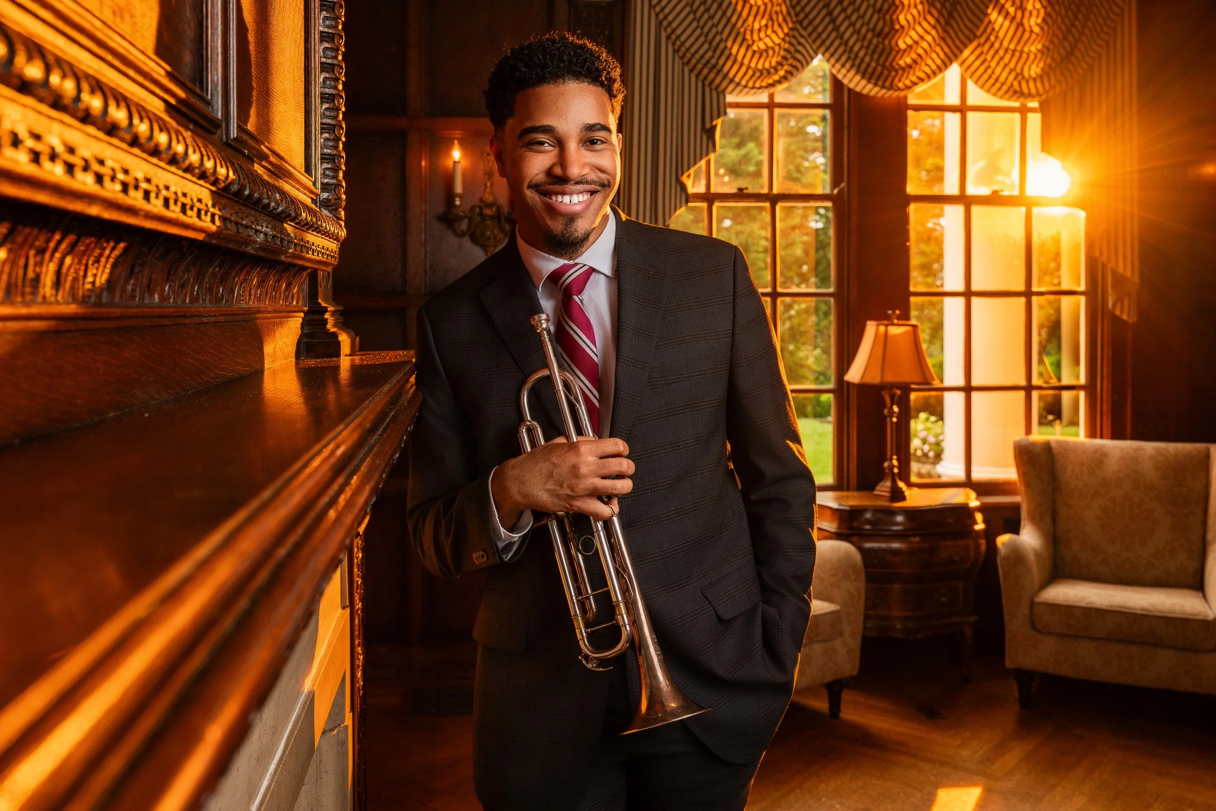 A man in a suit holding a trumpet, smiling, standing in a warm, wood-paneled room with large windows and sunset light, traditional curtains, and vintage lamps.