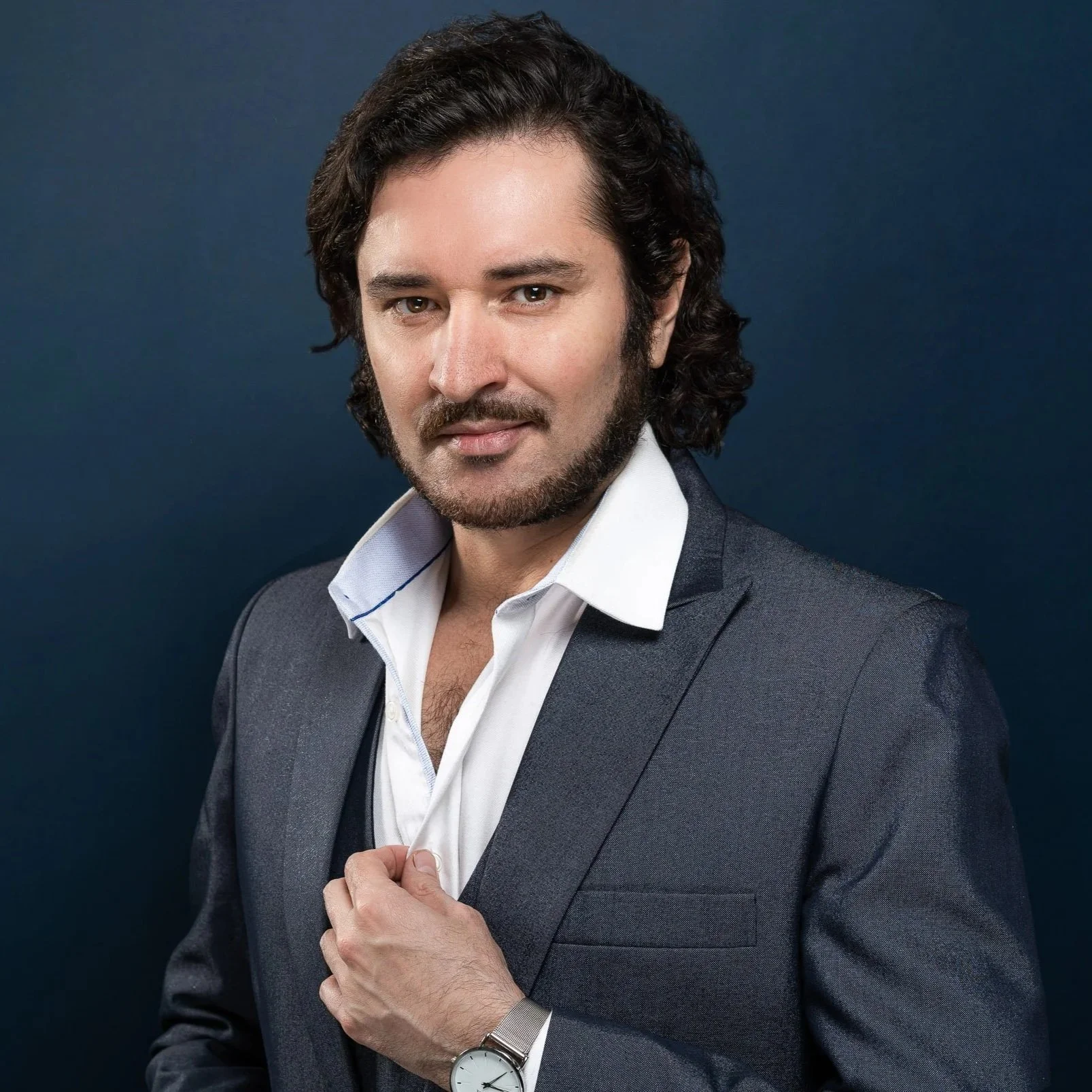 Man in a gray suit with a white shirt, adjusting his shirt with his right hand, wearing a silver wristwatch, against a dark blue background.