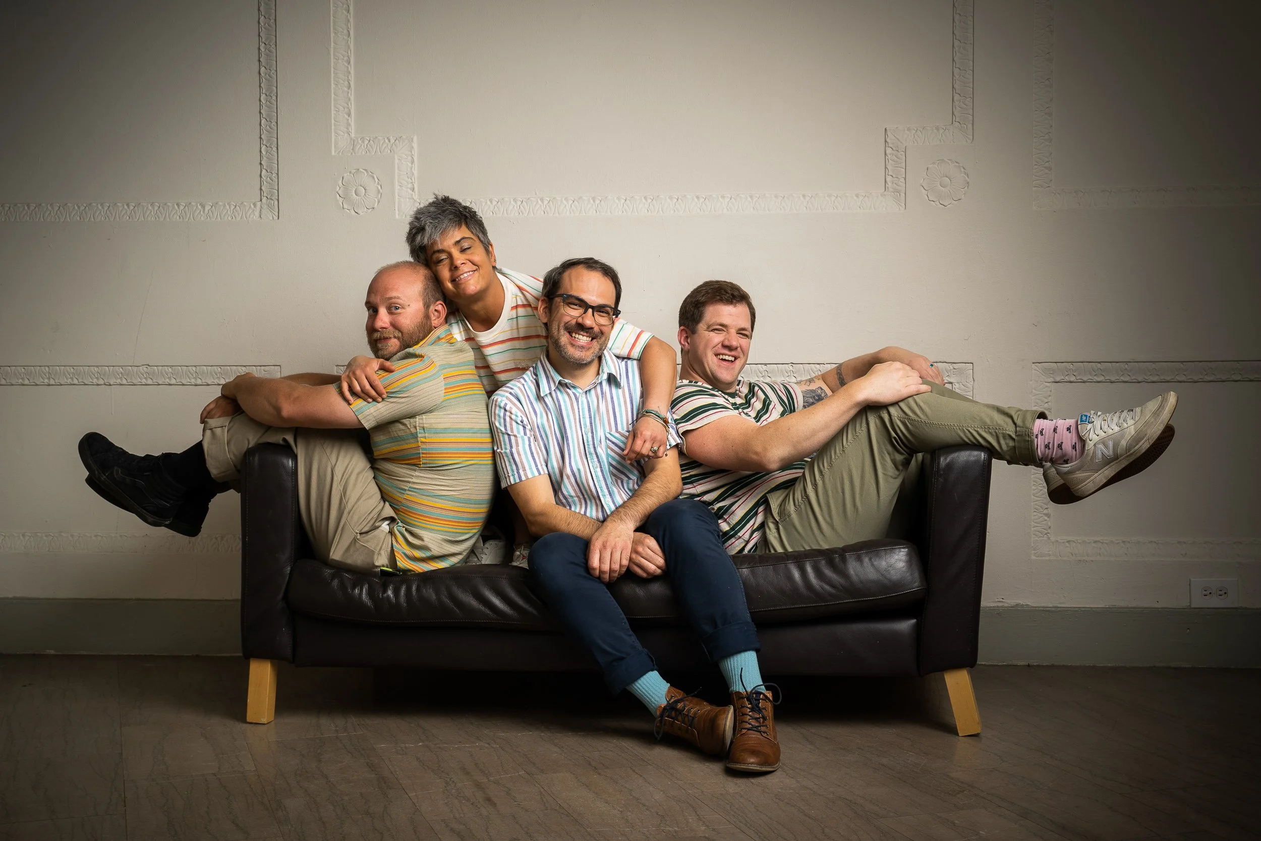 A group of five diverse friends sitting closely and happily on a dark brown sofa in a plain room with white decorated walls. They are dressed casually, laughing, and enjoying each other's company.
