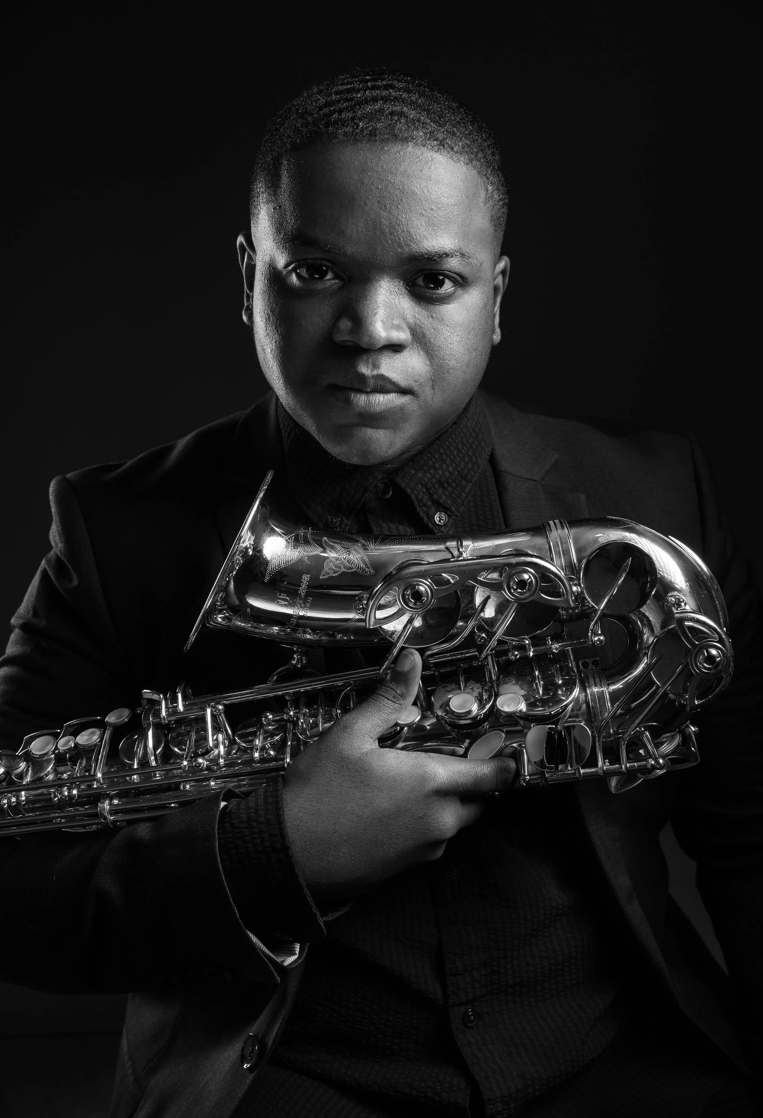 Black and white portrait of a person holding a saxophone, wearing a suit, with a serious expression, against a dark background.