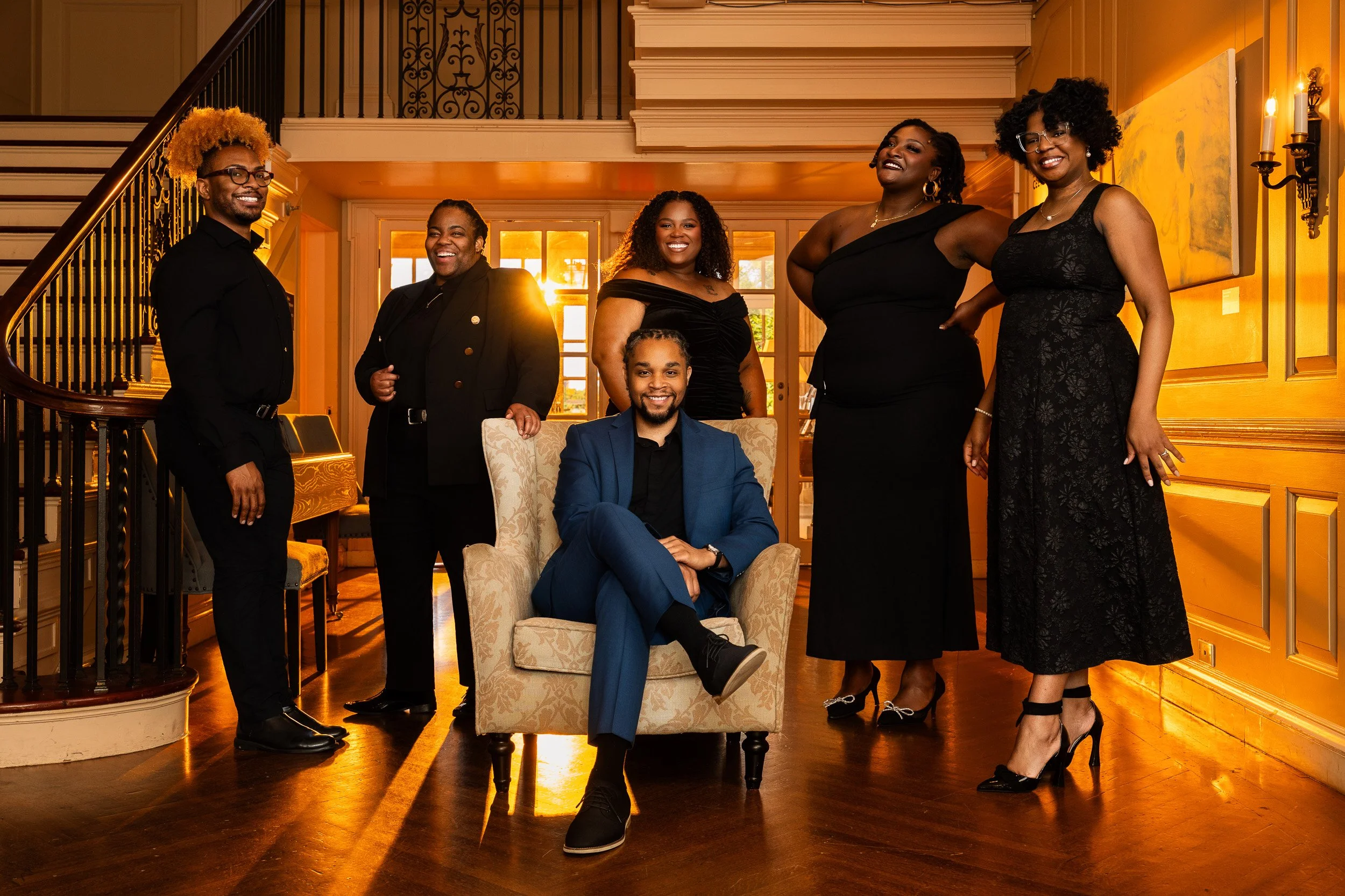 Group of seven diverse people, five women and two men, dressed in formal attire, posing in a warmly lit, elegant interior with wooden floors and staircase, smiling for the camera.
