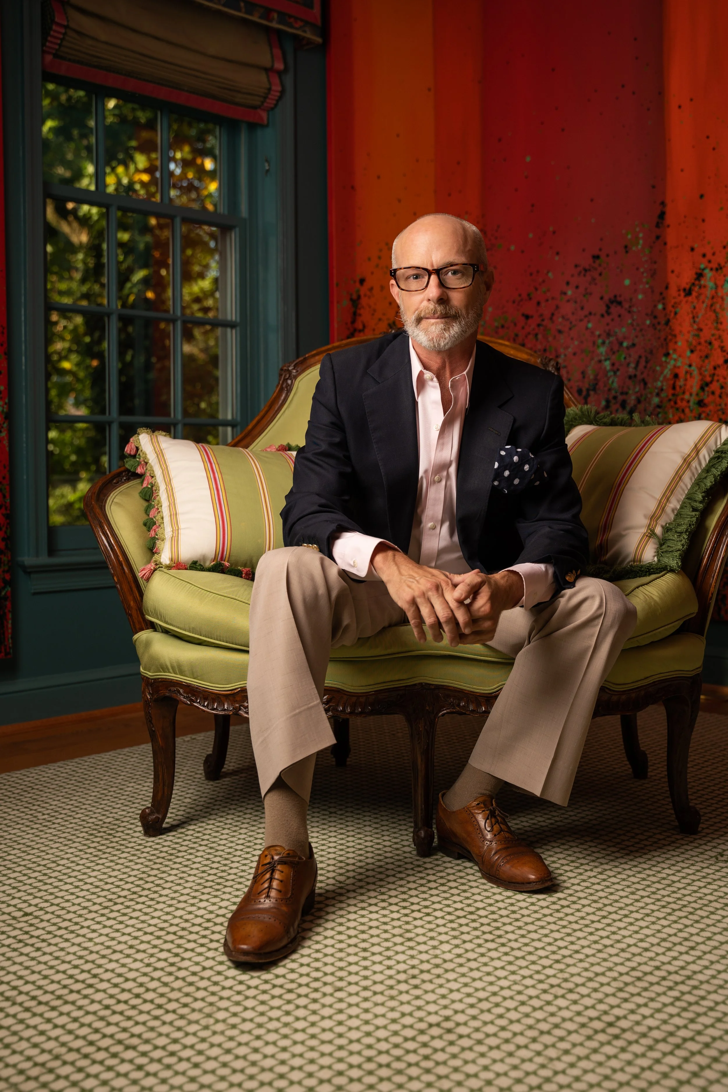 Bald man with glasses, beard, light pink shirt, and dark blazer sitting on a vintage sofa in front of a colorful abstract wall and window with greenery outside.