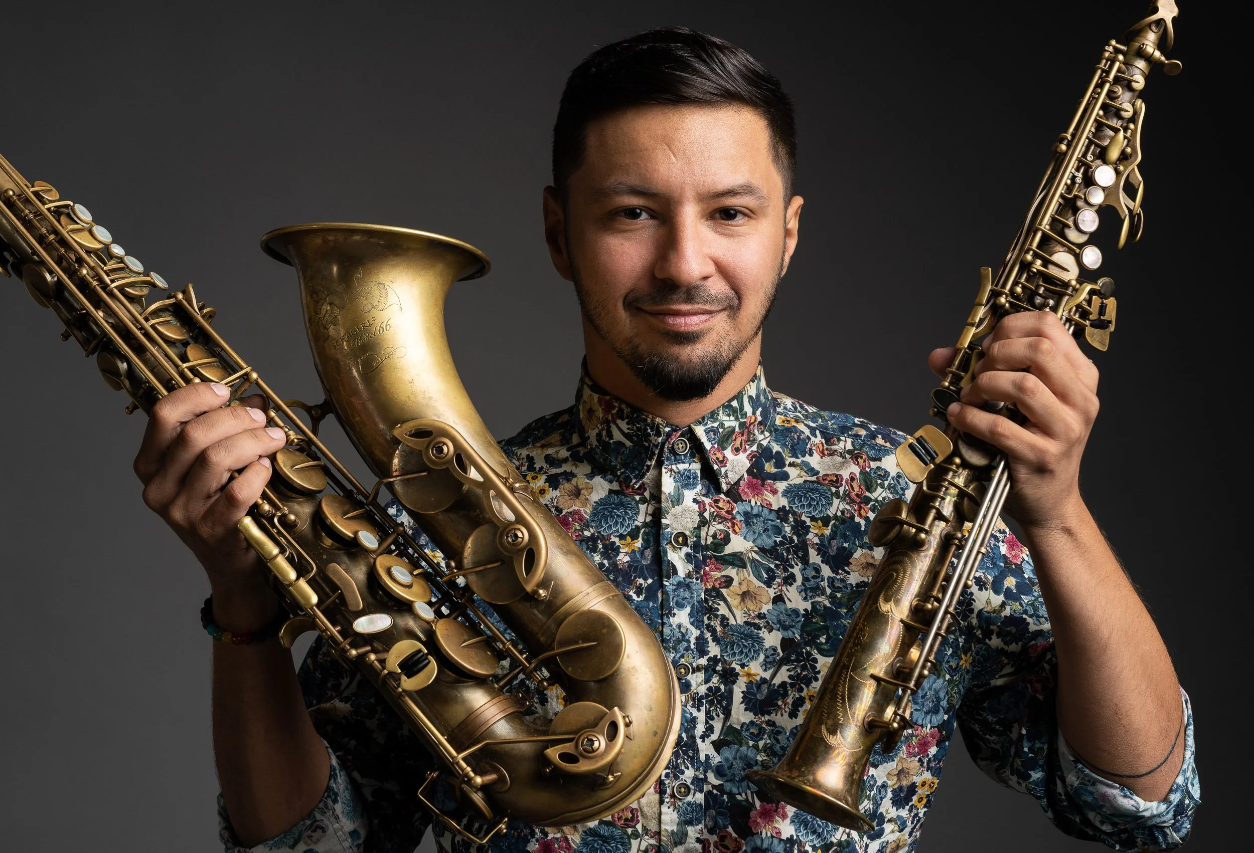 A young man with dark hair and a beard holding two saxophones in front of a dark gray background. He is wearing a colorful floral shirt and smiling slightly.
