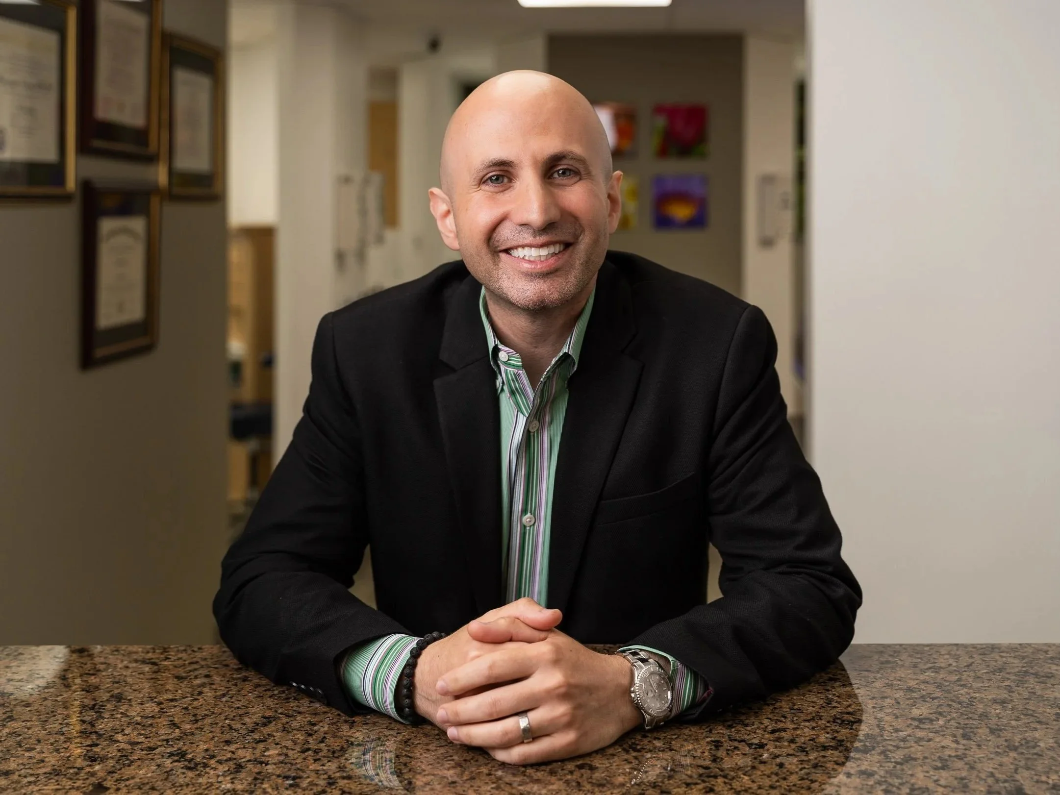 A smiling man with a bald head, wearing a black blazer over a green striped shirt, sitting at a granite countertop in an office or lobby area with art on the wall.