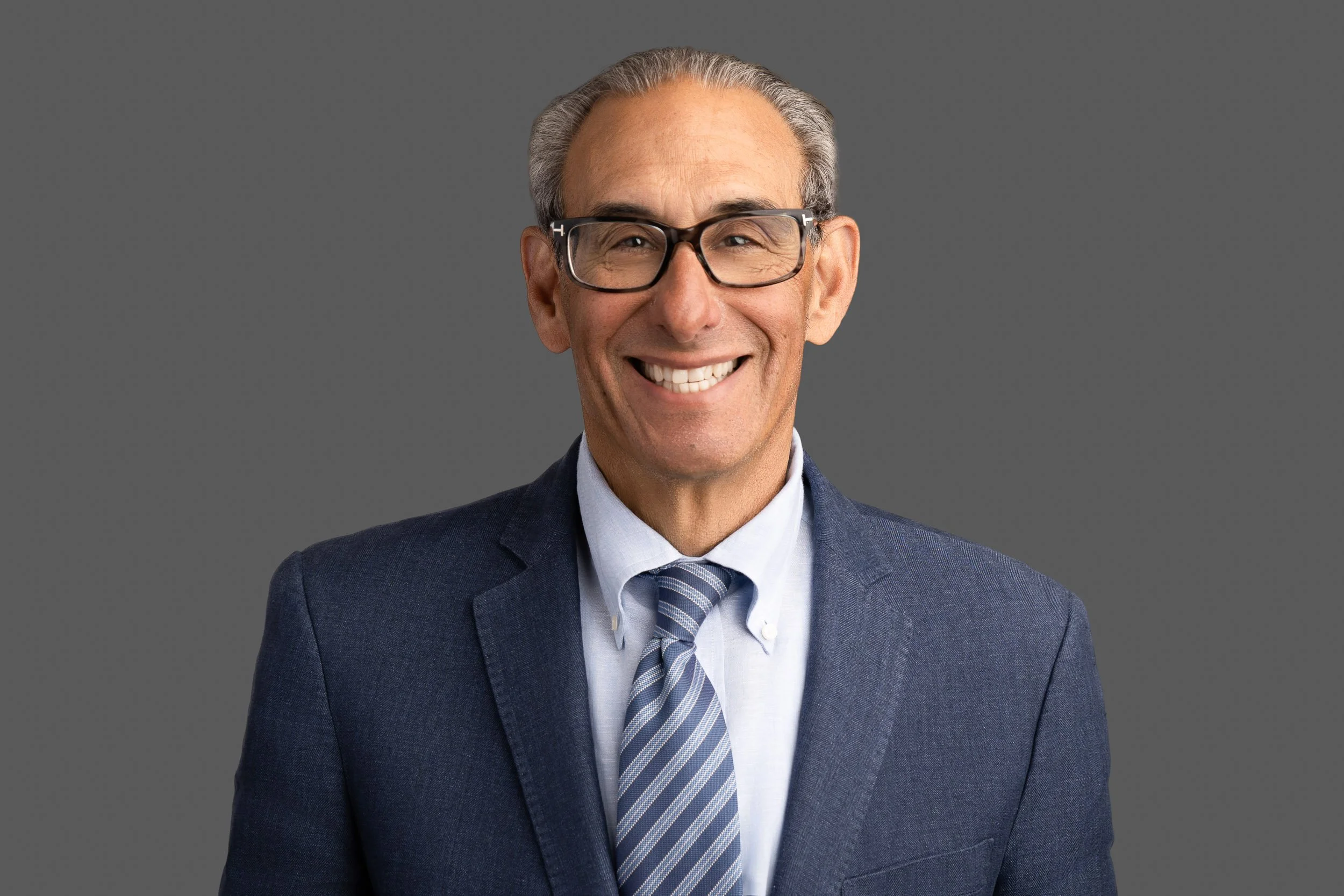 A smiling man with gray hair and glasses dressed in a navy blue suit, white shirt, and striped tie, standing against a plain gray background.