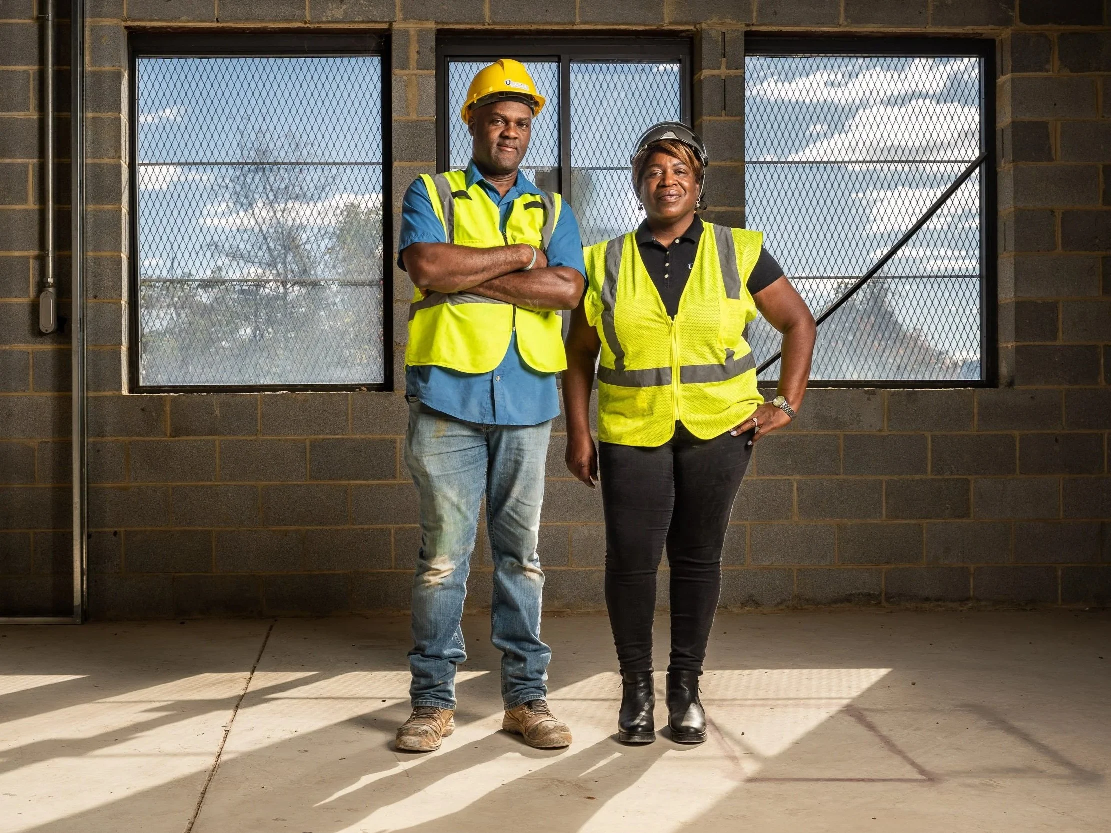 Two construction workers, one man and one woman, standing in front of a window with a wire mesh grate. Both are wearing yellow safety vests and helmets; the man has a blue shirt and jeans, while the woman wears a black polo shirt and black pants. They are posing confidently outdoors.