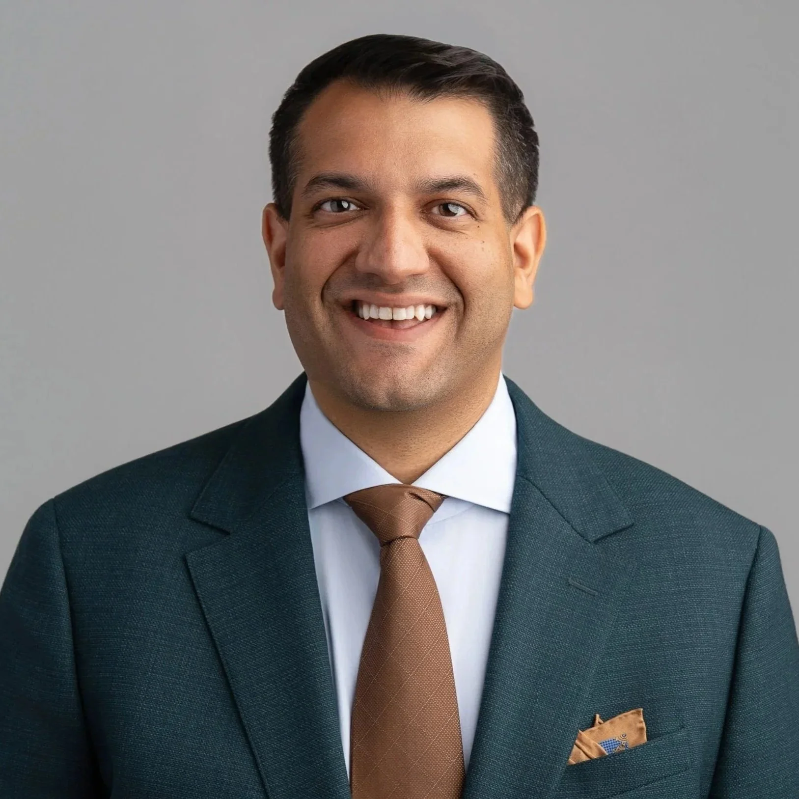 A smiling man wearing a dark green suit, white shirt, brown tie, and a matching brown pocket square, posed against a plain gray background.