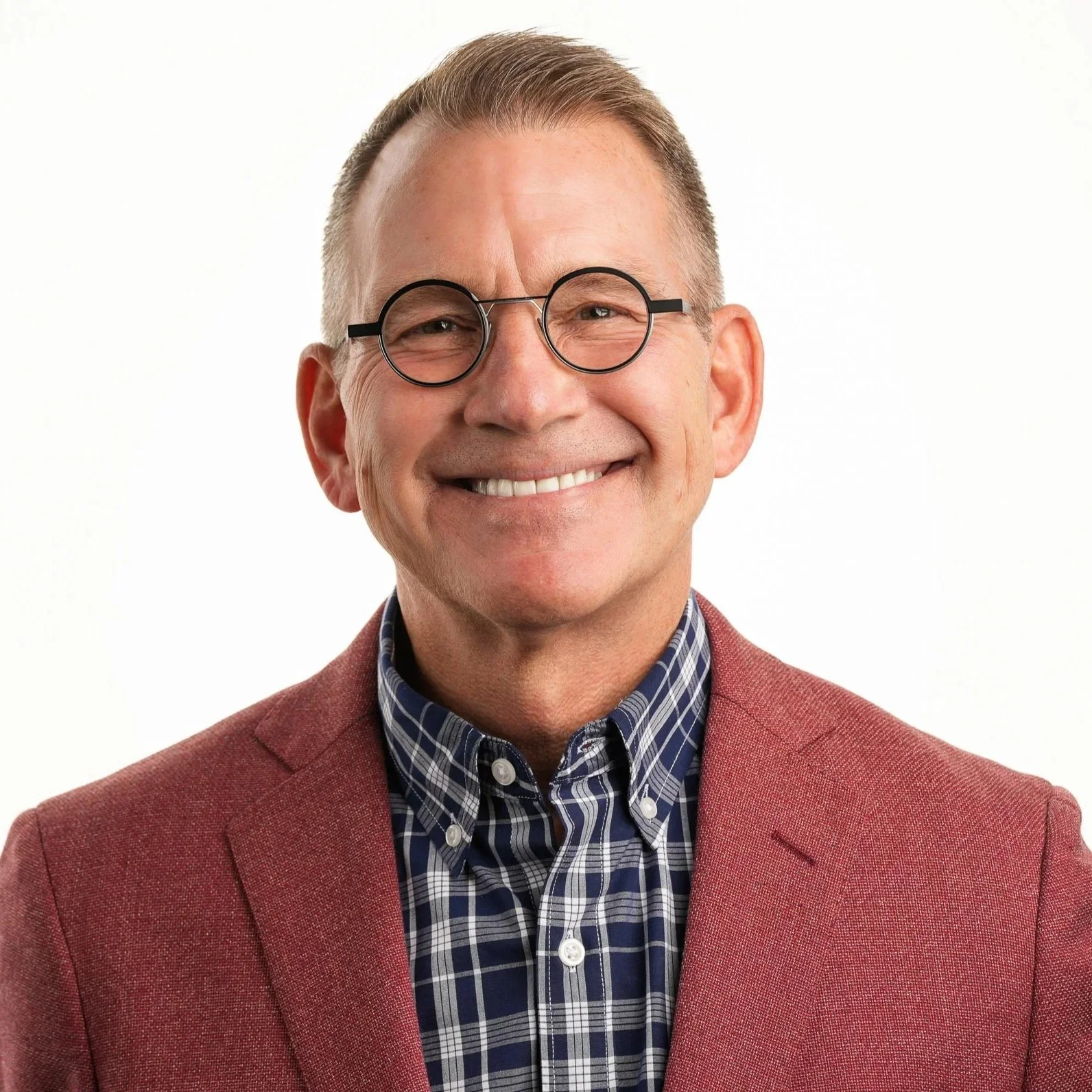 A smiling middle-aged man with short, light brown hair, wearing round black glasses, a plaid shirt, and a red blazer, against a plain white background.