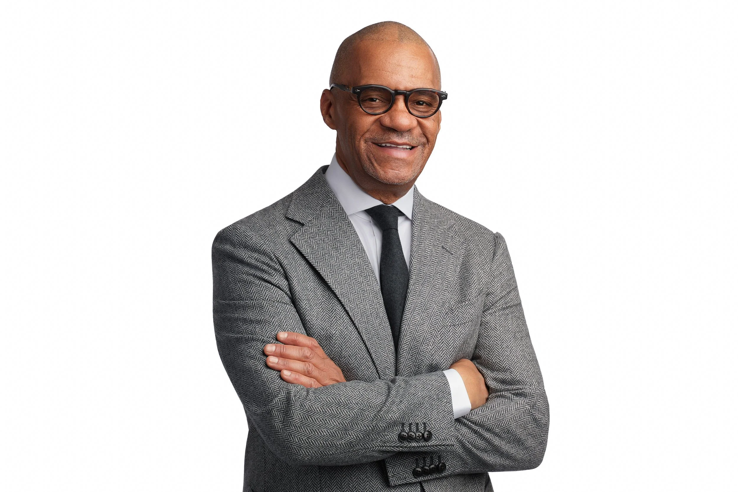 A middle-aged African American man wearing glasses, a gray checkered suit, white shirt, and black tie, smiling with arms crossed against a white background.