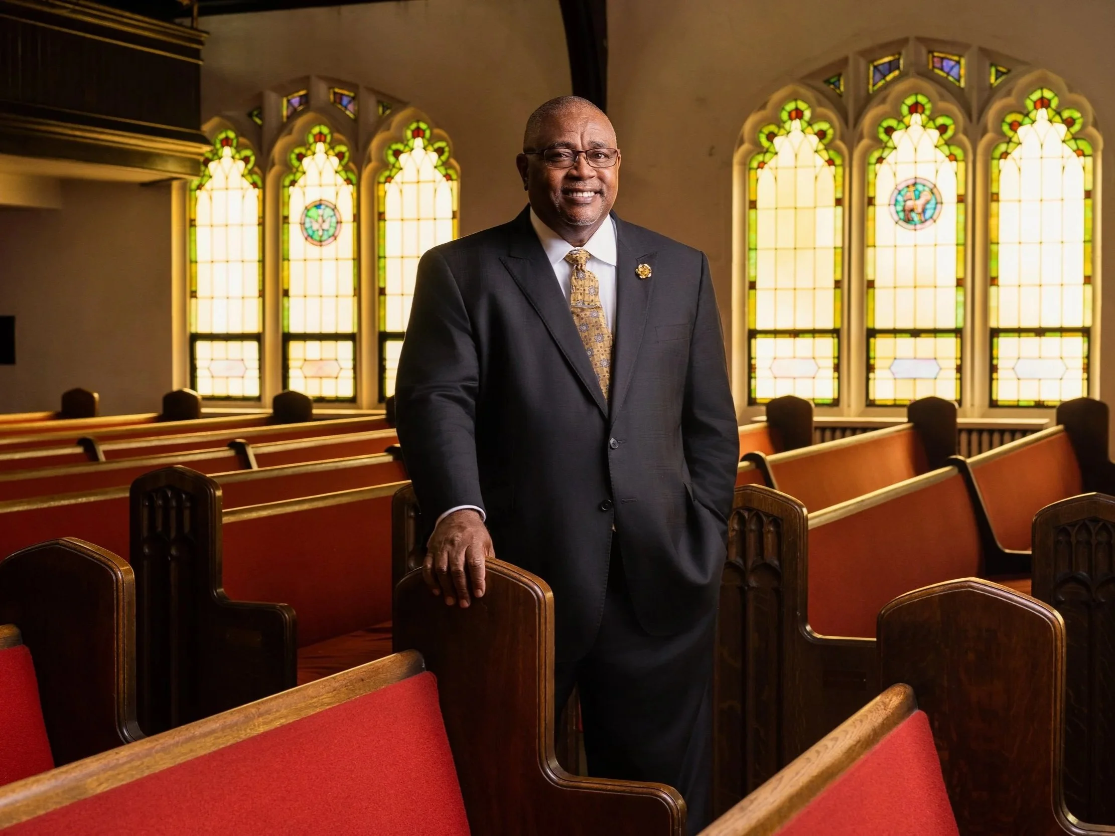 An older man in a suit and tie standing inside a church, with colorful stained glass windows behind him and wooden pews in the foreground.