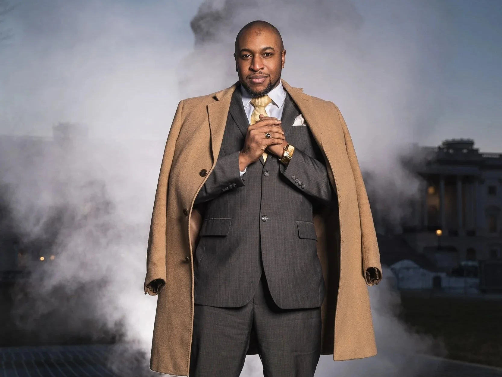 A confident man in a dark gray suit with a white shirt and yellow tie, wearing a tan overcoat draped over his shoulders, standing outdoors with smoke and buildings in the background.