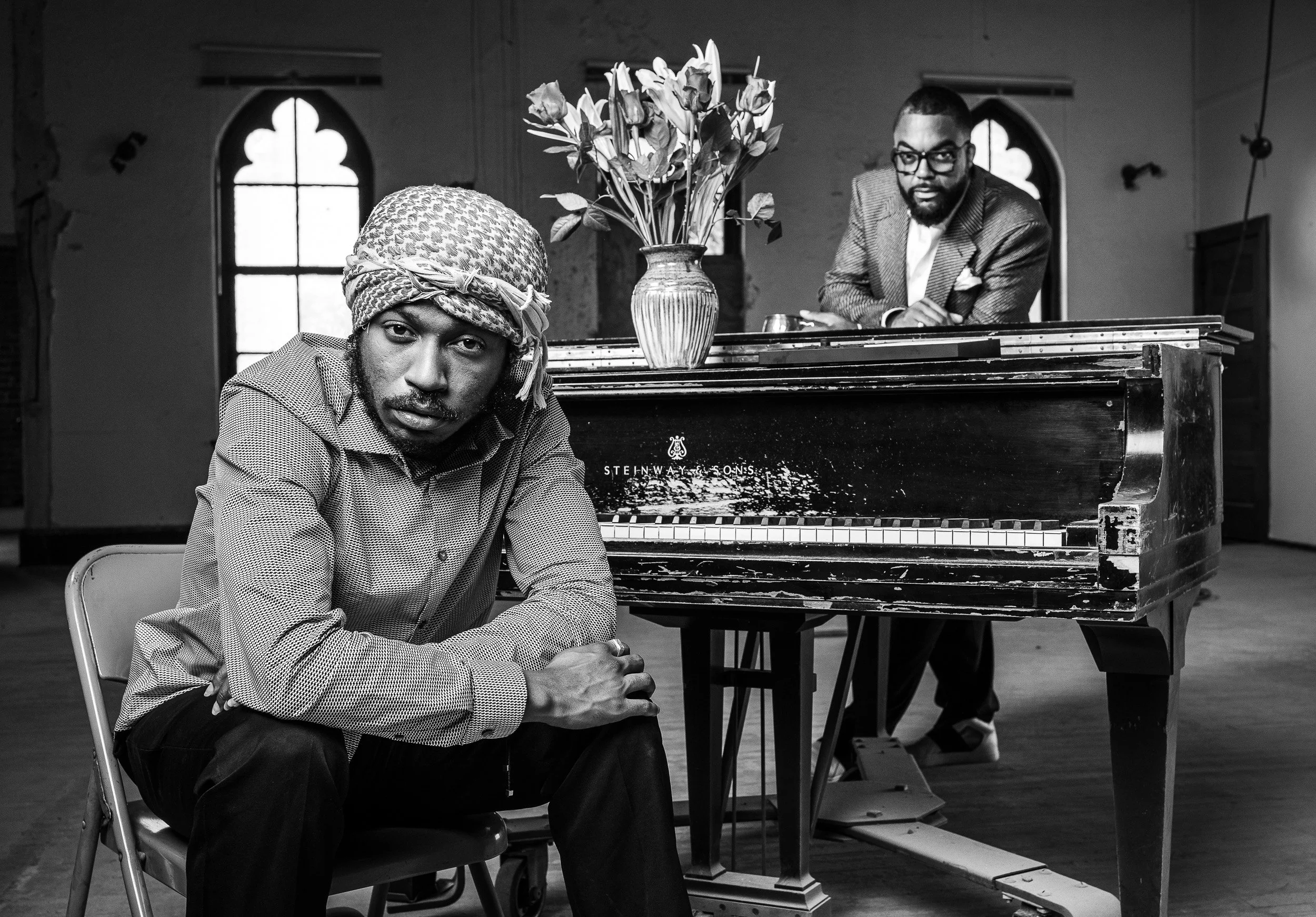 Black and white photo of two men in a room with gothic windows; one man is sitting in a chair at a table, wearing a patterned shirt and a headscarf, looking at the camera, while a man in a suit stands behind a piano, resting his arms on it, with a serious expression. There is a large vase with flowers on the piano.