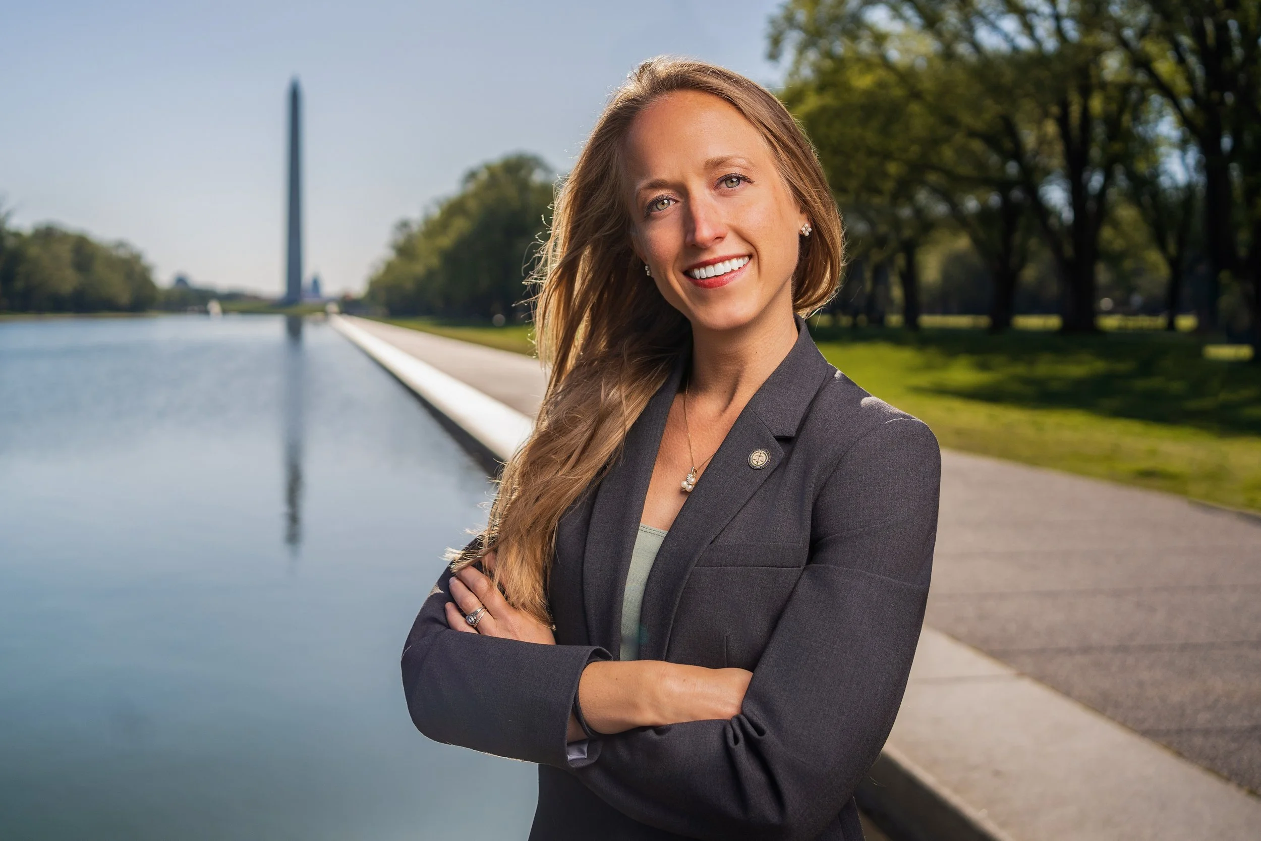 A woman with long blonde hair, wearing a dark blazer with a lapel pin, standing outdoors near water with a path and trees, smiling with crossed arms.