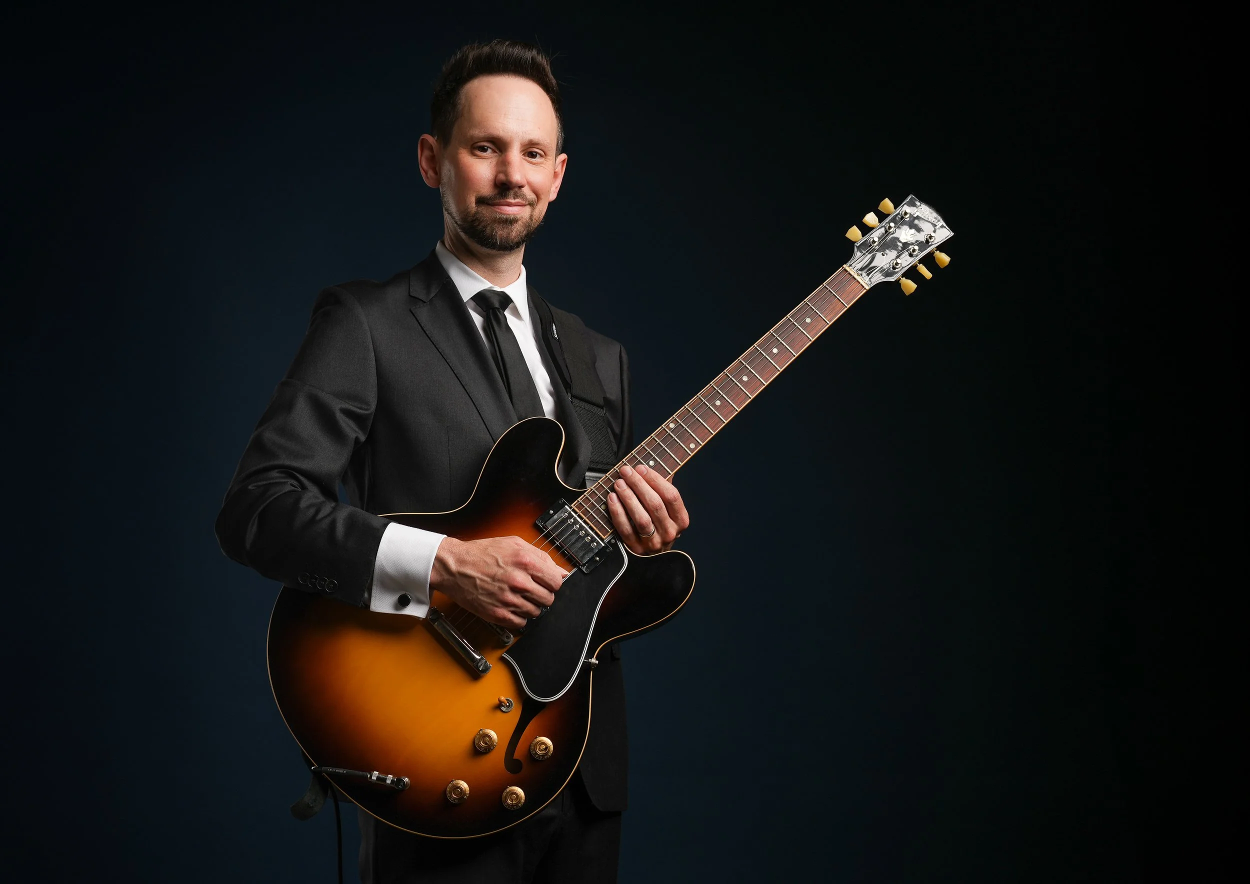 A man in a formal black suit and tie holding an electric guitar against a dark background.