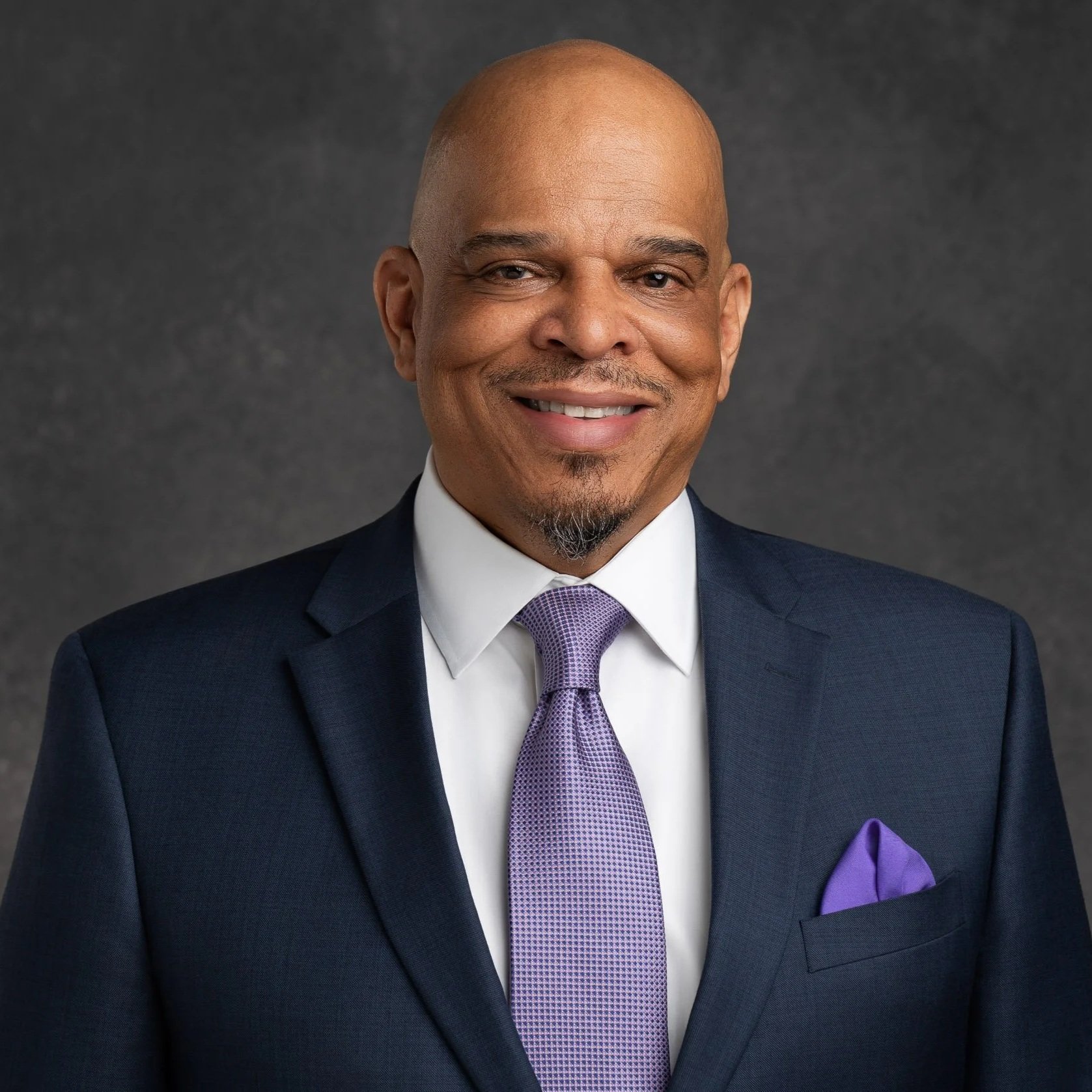 Headshot of a smiling man wearing a navy suit, white shirt, purple tie, and a matching pocket square, against a dark gray background.
