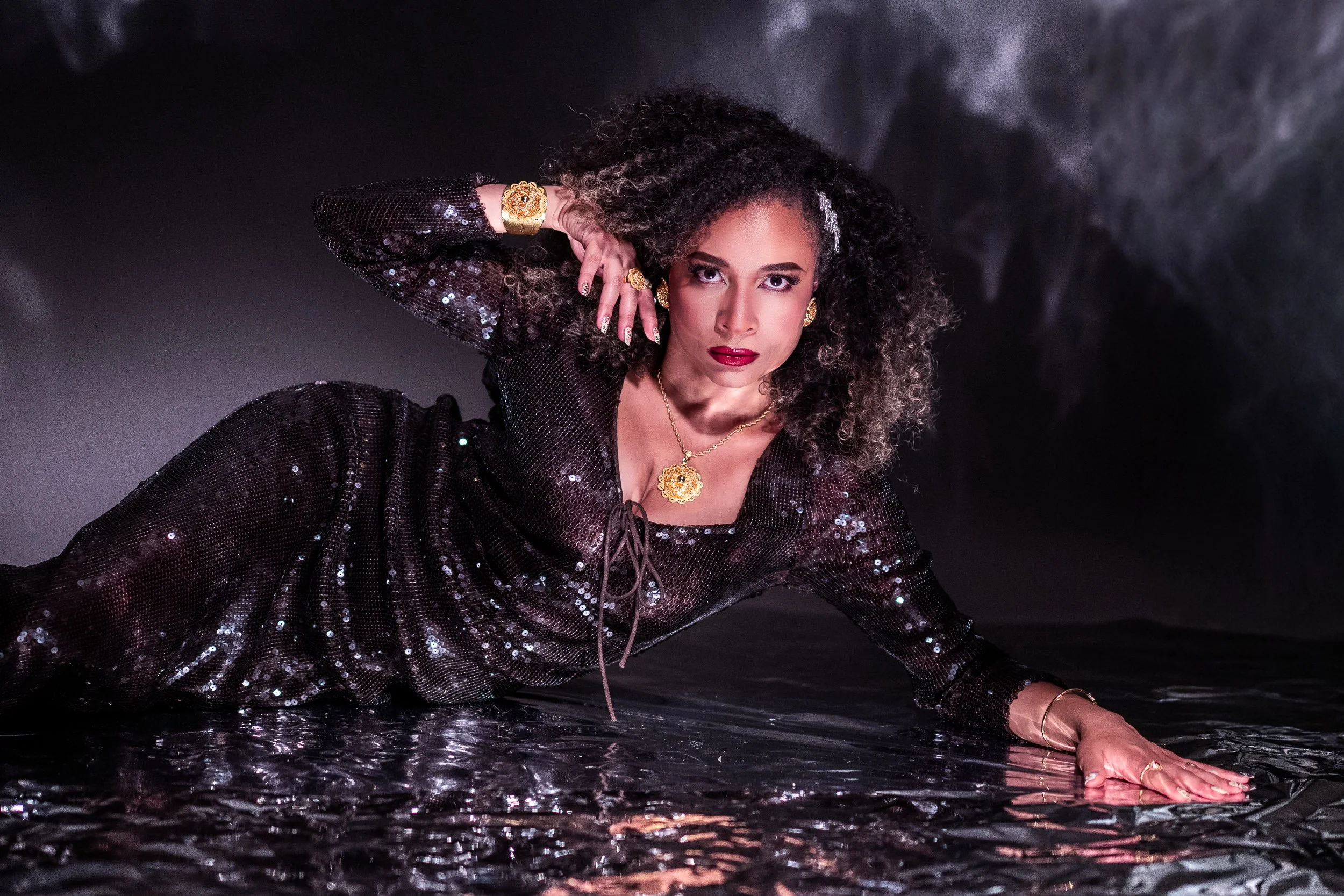 A woman with curly hair posing against a dark smoky background, wearing a black sequined outfit and gold jewelry.
