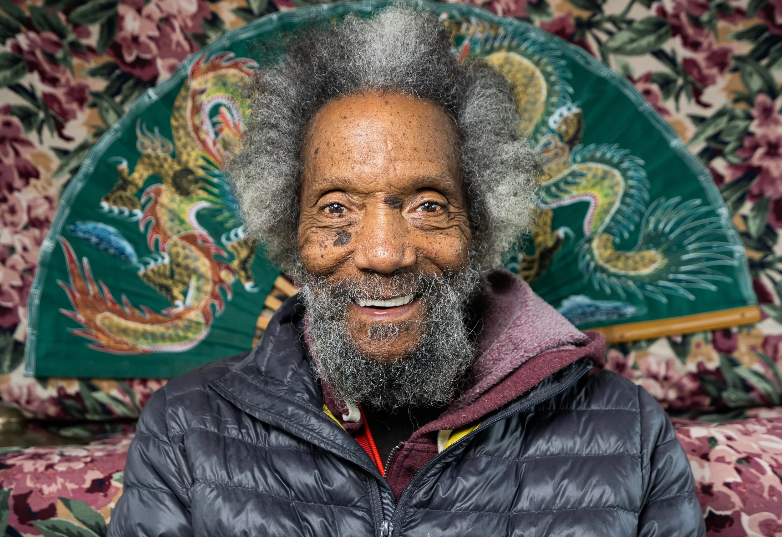 Older man with gray, curly hair and beard, smiling, lying on a colorful floral-patterned blanket, wearing a black puffy jacket.