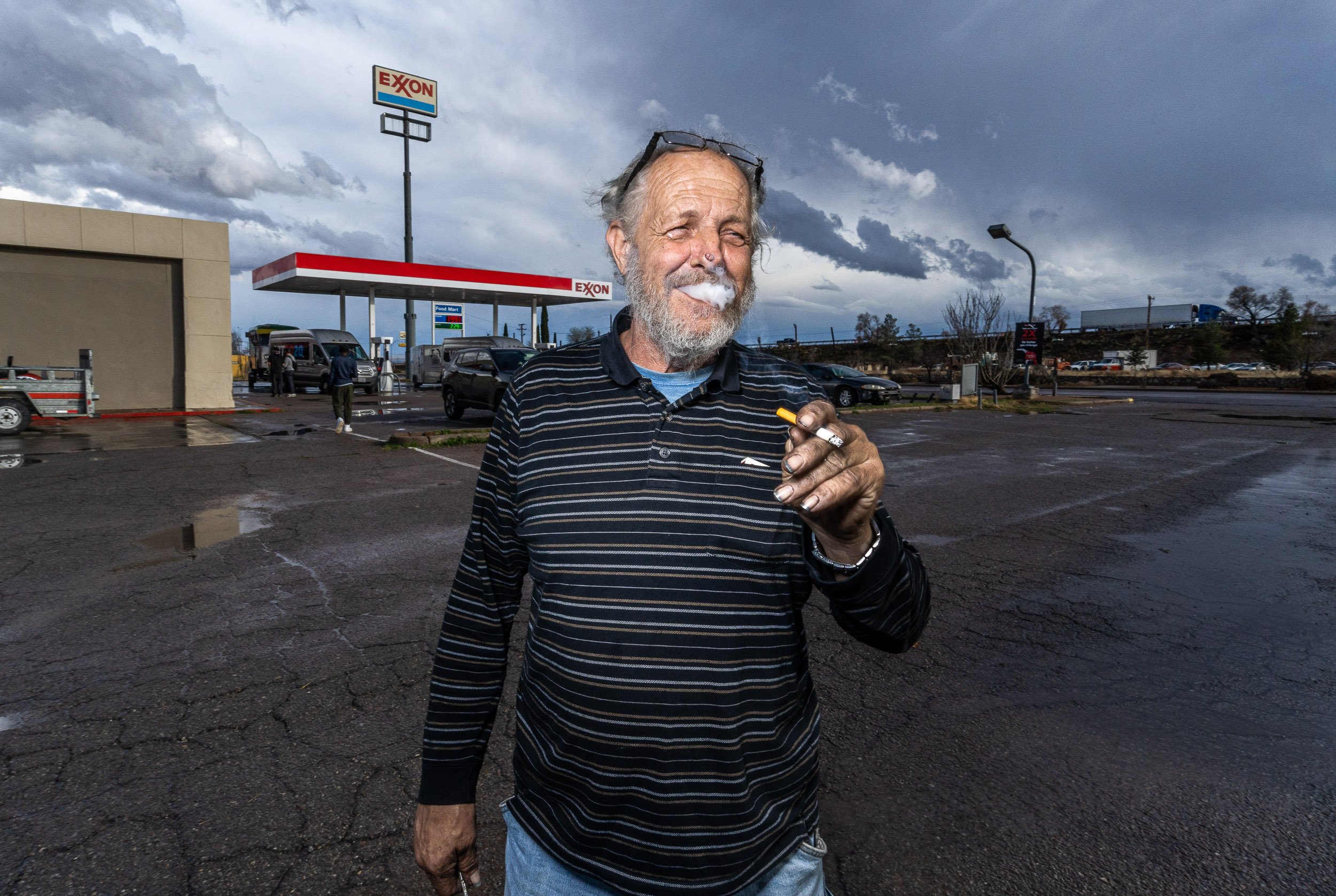 An elderly man with a white beard and glasses on his head, smoking and smiling while standing in a parking lot near an Exxon gas station on a cloudy day.