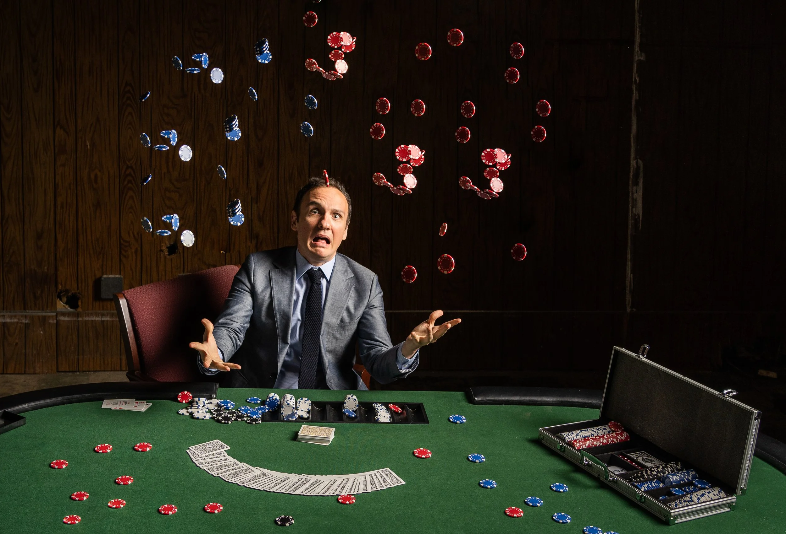 A man in a gray suit with a surprised expression sits at a poker table with red, black, blue, and white poker chips scattered and in stacks, along with playing cards and a wallet with additional chips. Chips are flying in the air above him, as if he has just thrown or is throwing chips.