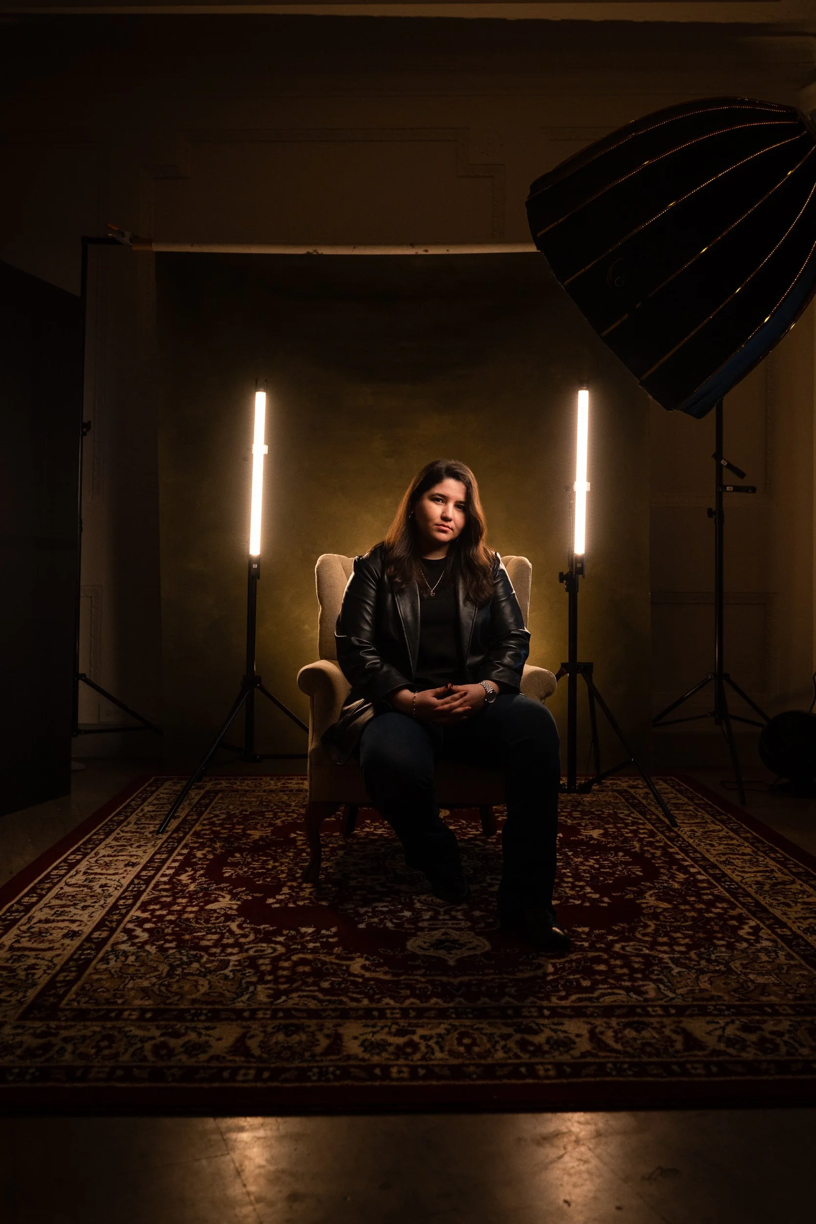 A woman with brown hair, wearing a black leather jacket and dark clothing, sitting on a beige armchair in a photo studio with studio lights and dark background.