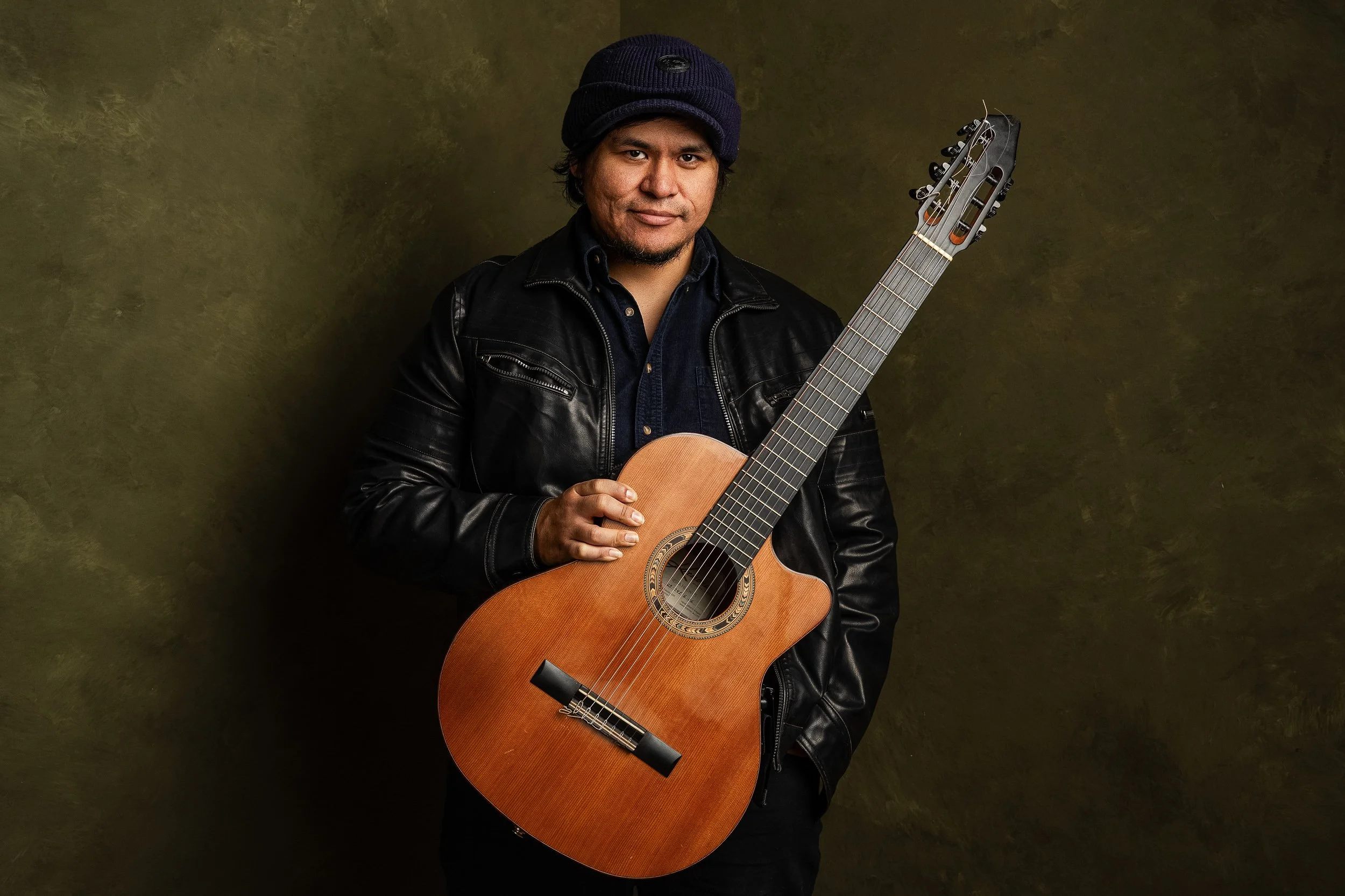 A man with dark hair and a beard wearing a black jacket and a knit cap, holding an acoustic guitar, standing against a textured olive green background.