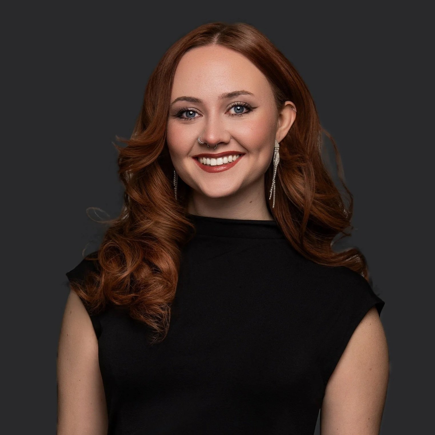 Portrait of a smiling young woman with long, wavy red hair, wearing a black top, silver earrings, and a septum piercing, against a dark background.
