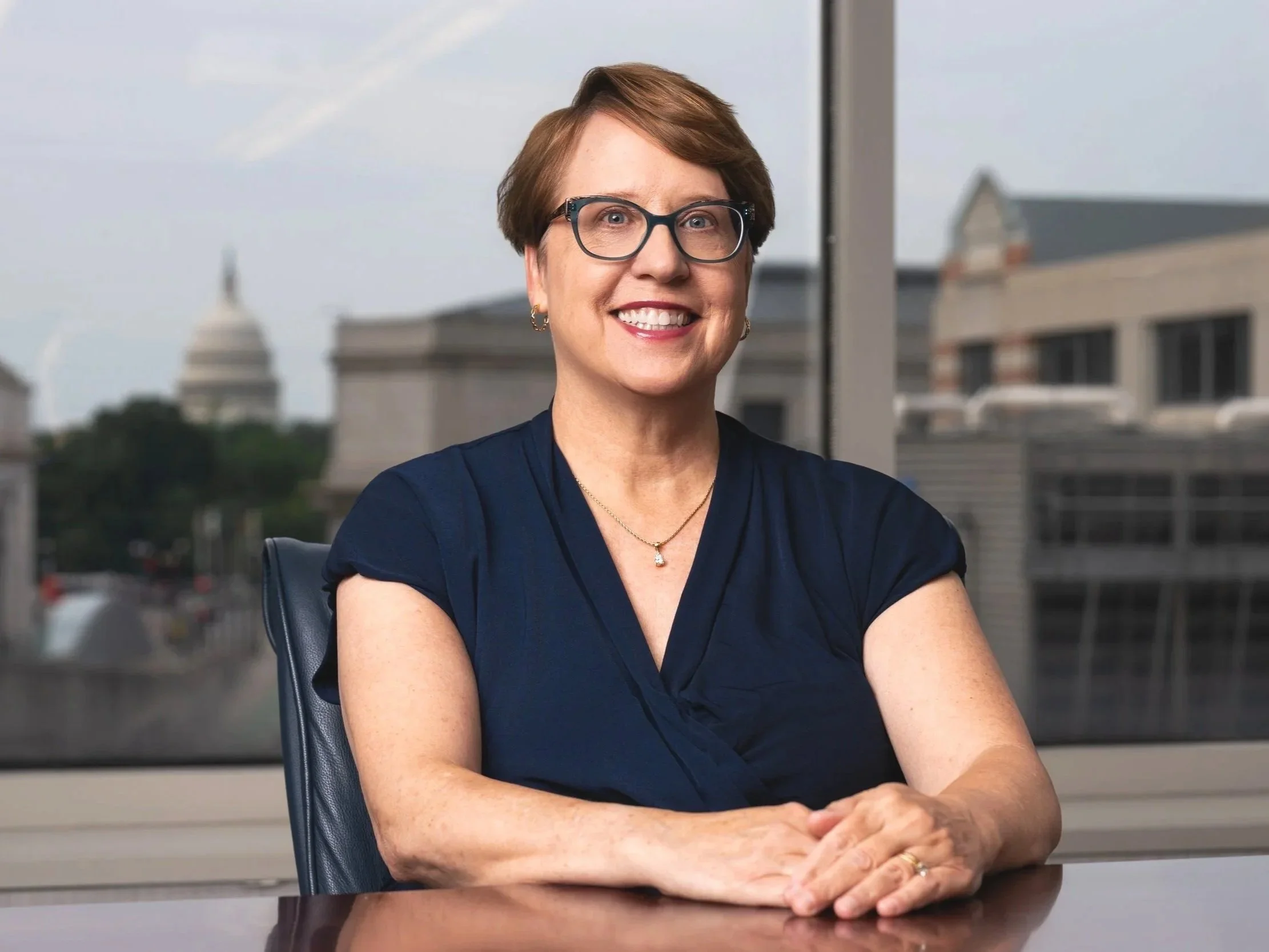 A woman with short brown hair, glasses, and earrings smiling in an office, with a cityscape view through the window behind her.