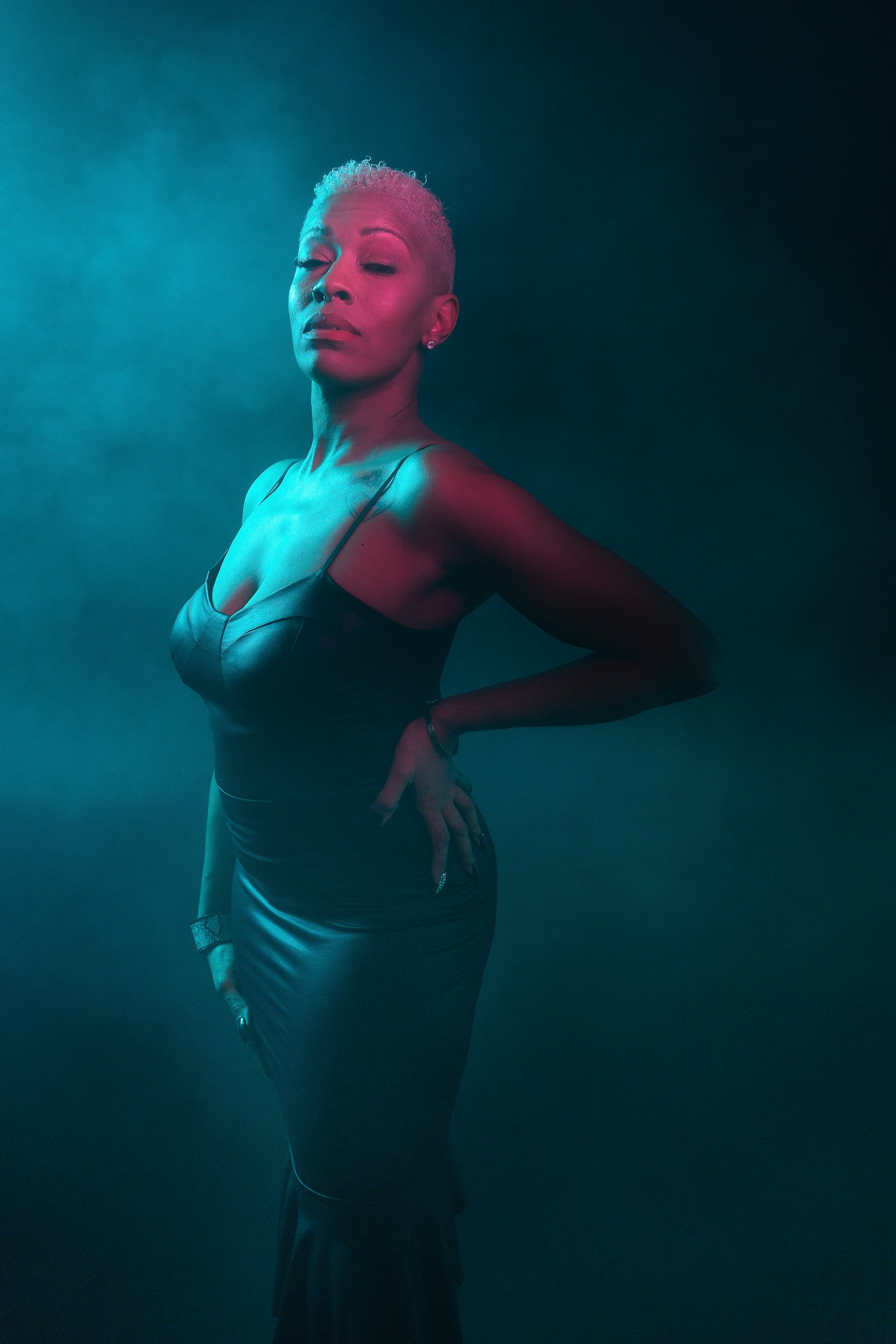 A woman with short blonde hair, wearing a sleeveless black dress, standing against a gradient background with blue and pink lighting.