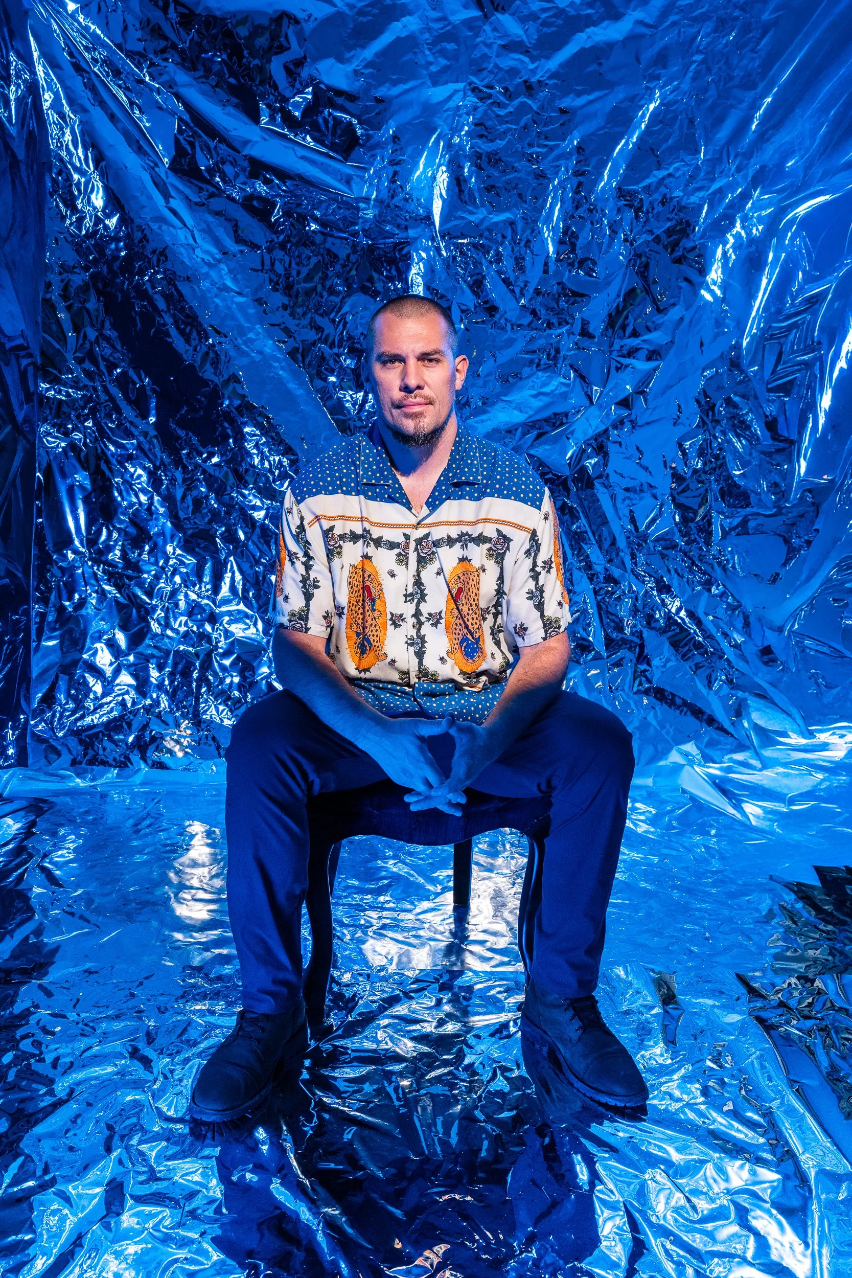 A man with a shaved head and goatee, wearing a patterned button-up shirt, sits on a black chair in front of a shiny, crinkled blue metallic backdrop.