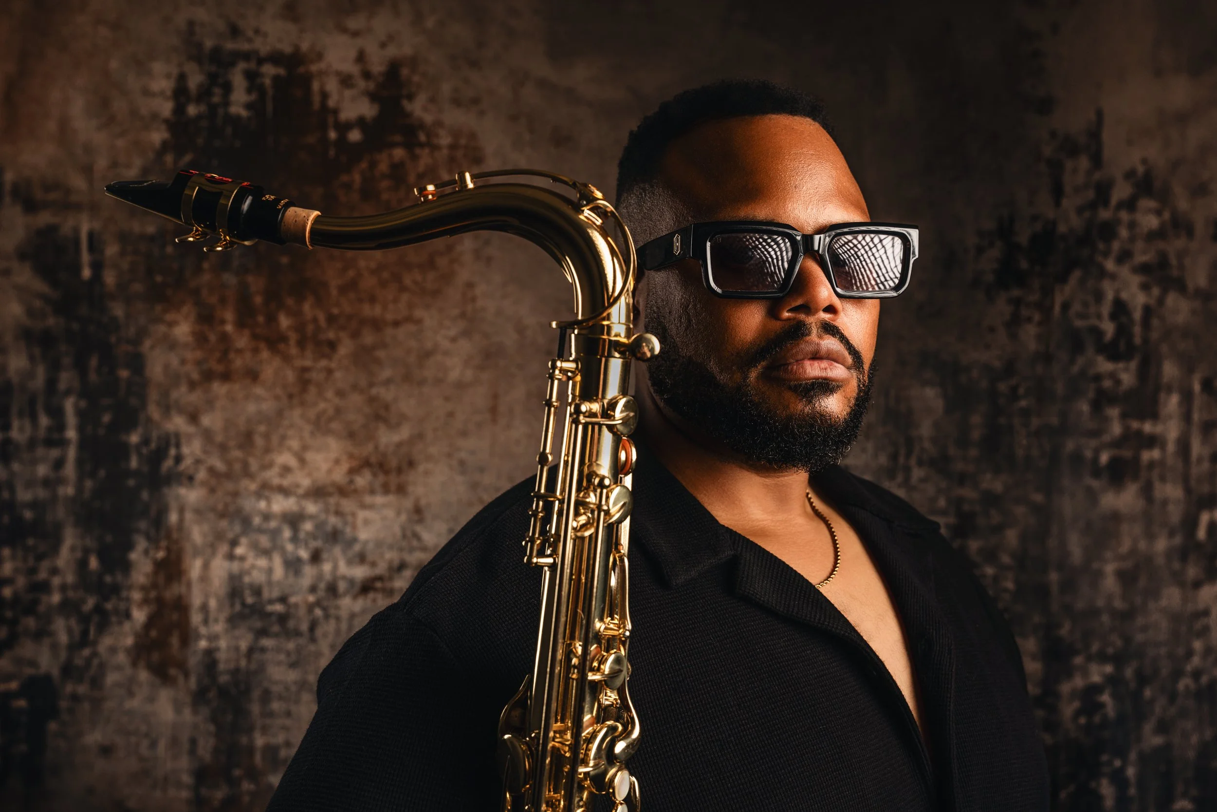 A man with dark skin, a beard, and short hair wearing black sunglasses, holding a gold saxophone on his shoulder, against a dark, textured background.