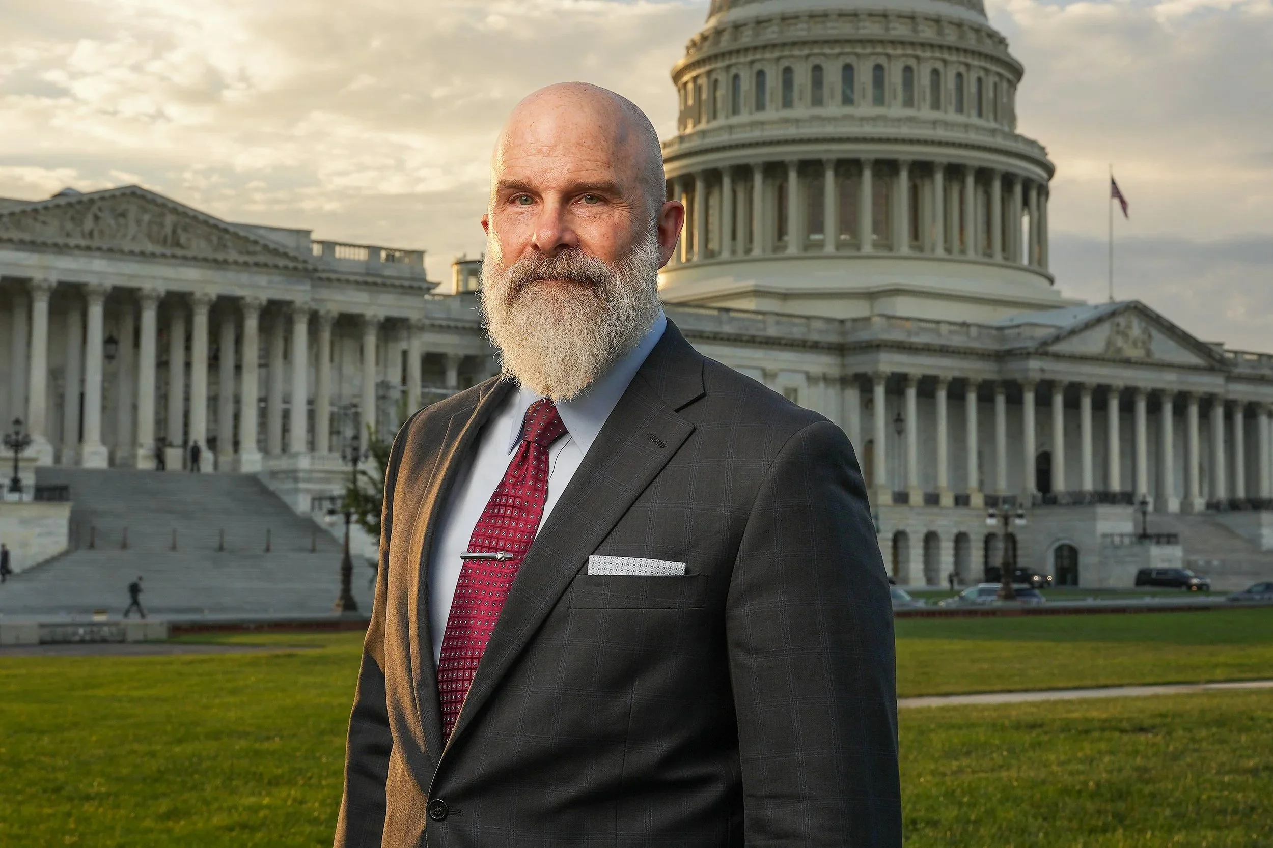 A man with a bald head and a long white beard wearing a gray plaid suit, white shirt, and red tie, standing outdoors in front of the U.S. Capitol building at sunset.