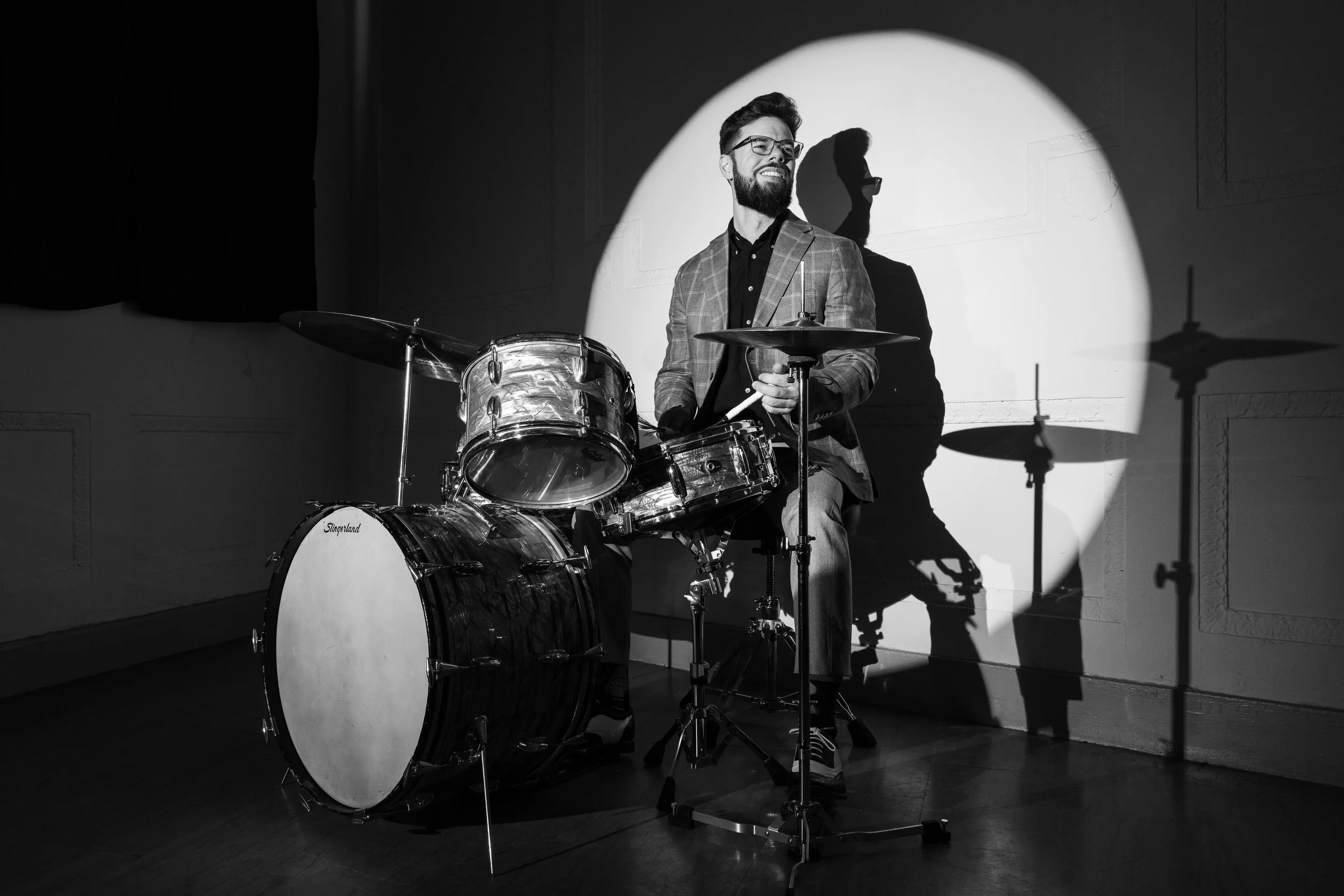 A man with glasses, a beard, and a plaid blazer playing drums under a spotlight in a dark room.