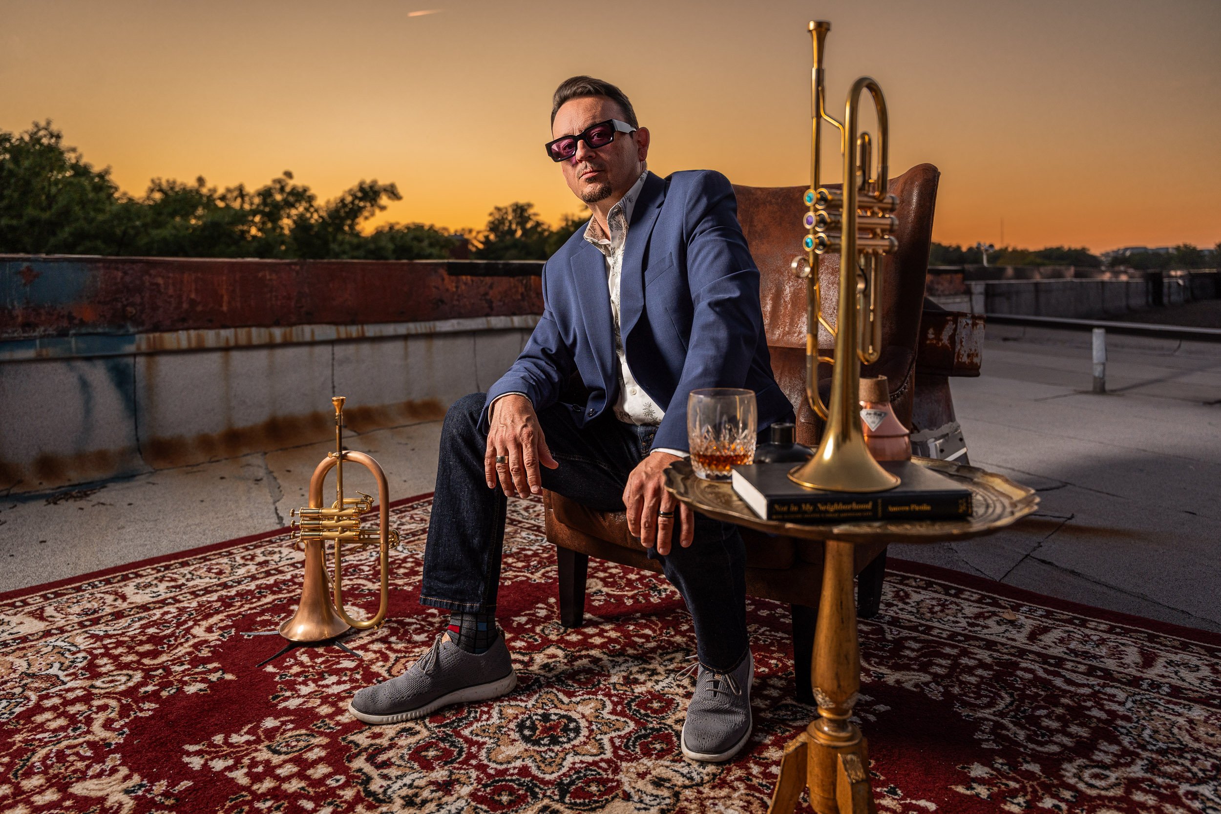 Man in a blue suit sitting on a rooftop terrace during sunset, with a trumpet and musical accessories around him, including a glass of whiskey, a book, and a pair of sunglasses.