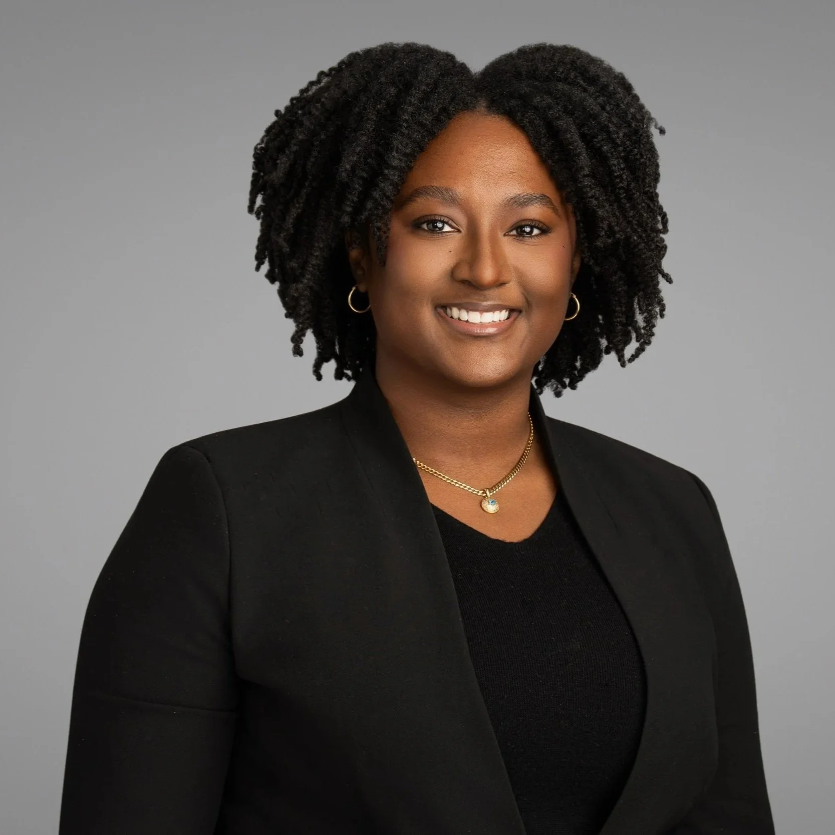 A portrait of a smiling Black woman with natural curly hair, wearing a black blazer and jewelry, against a gray background.