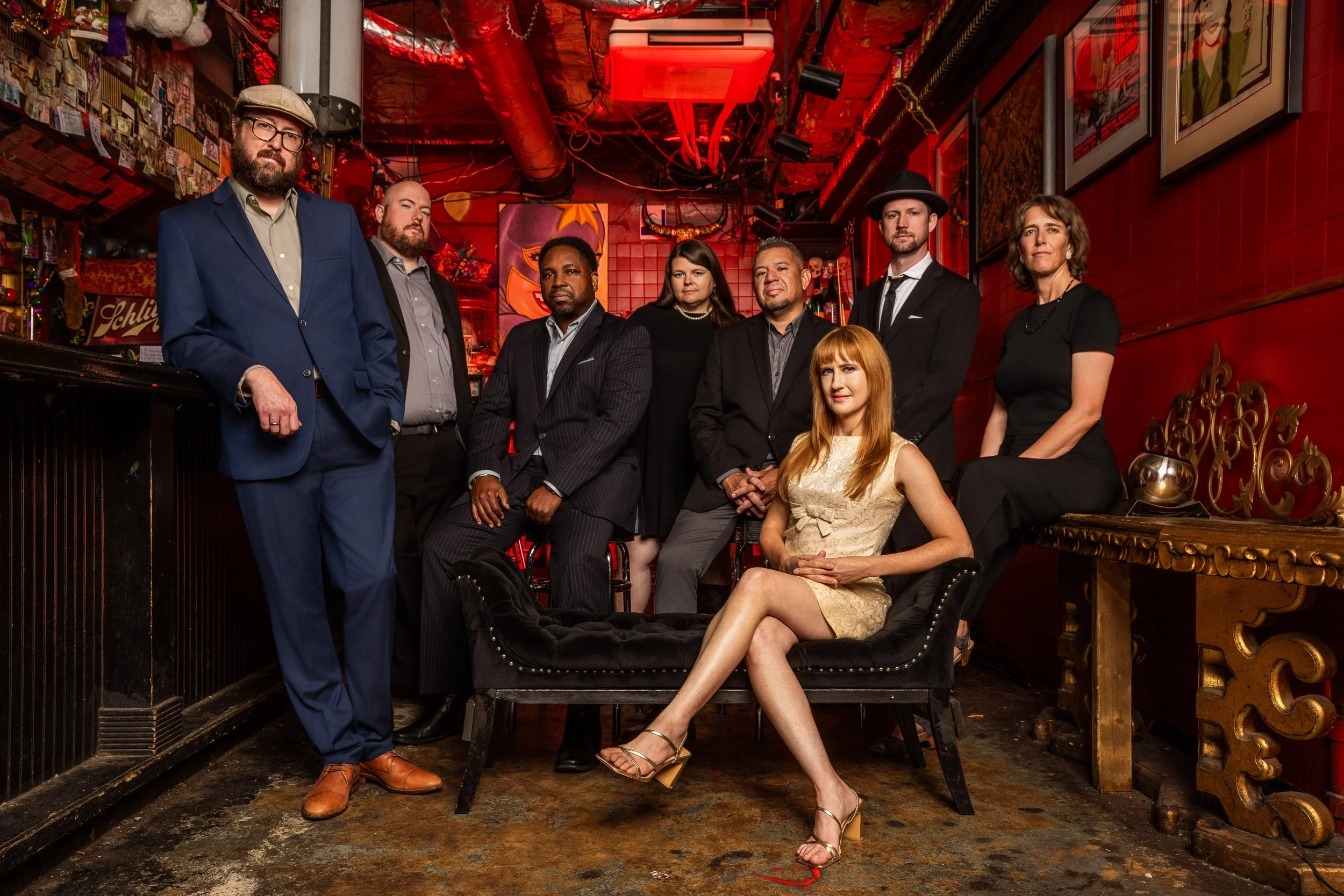 Group of nine diverse people in stylish clothing posing in a dimly lit, red-themed bar or lounge with artwork and movies posters on the walls.