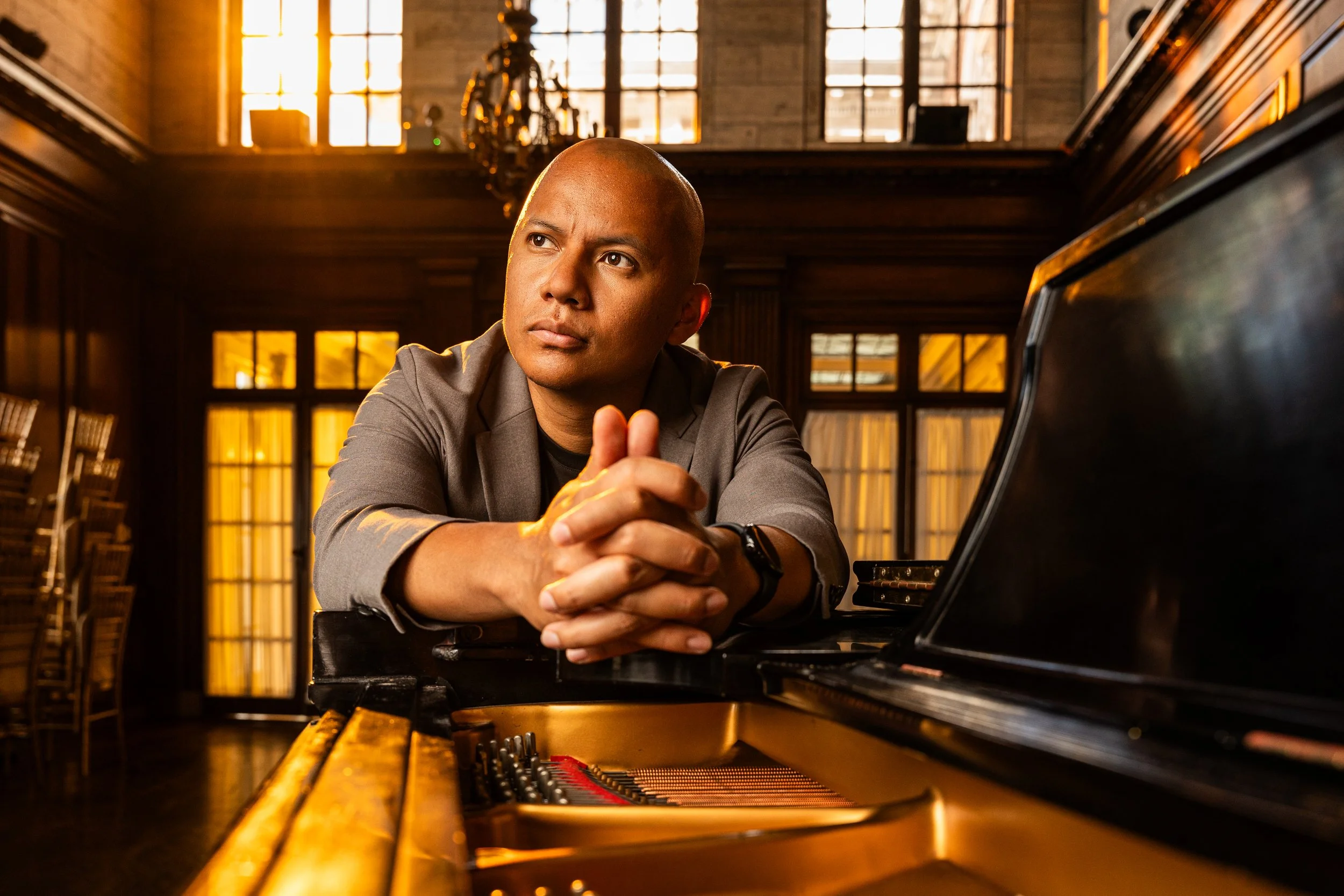 A man sitting at a grand piano in a well-lit room with large windows and wooden interior.
