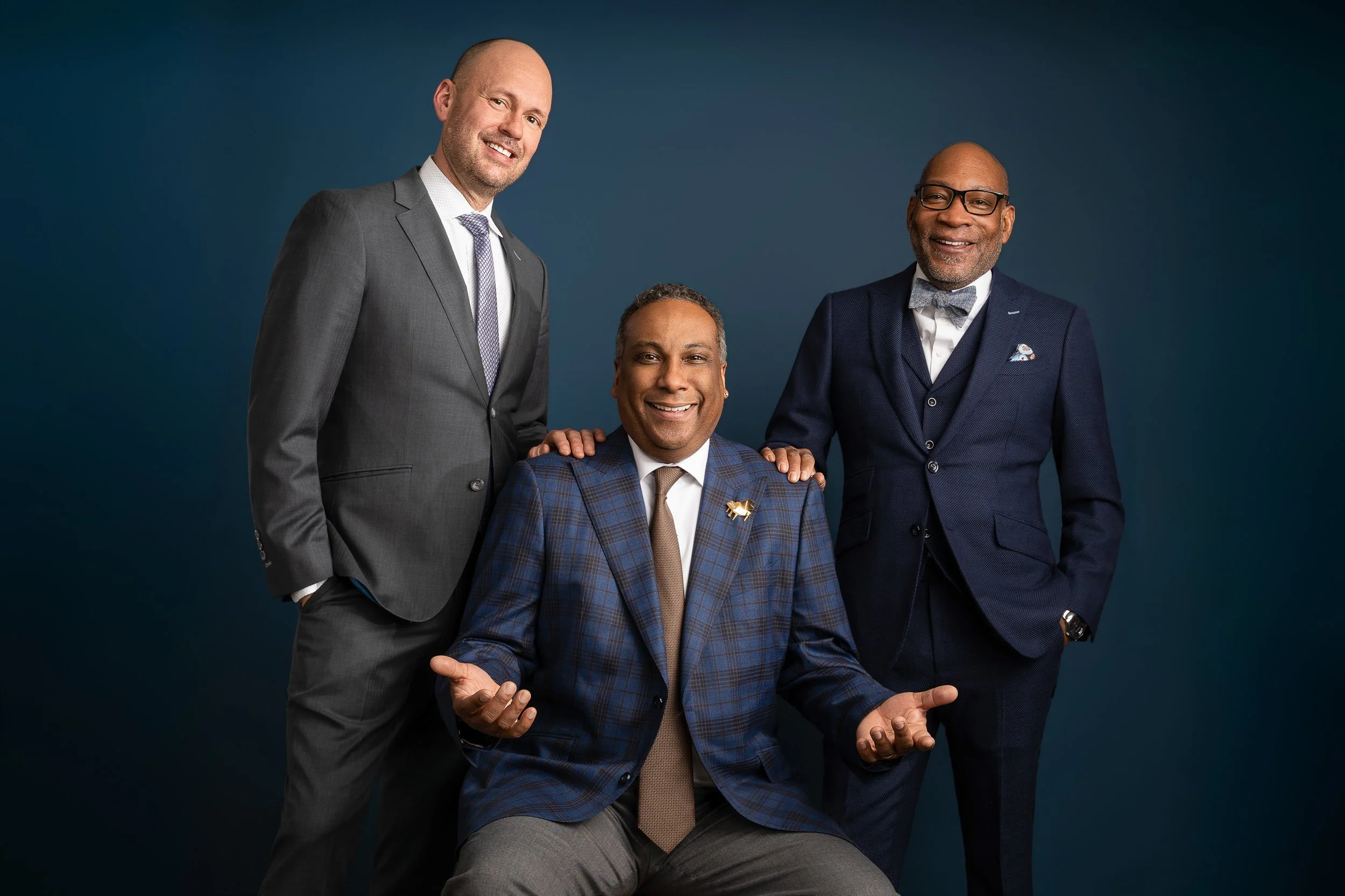 Three men dressed in suits, smiling, against a dark blue background. The man in the middle, seated, is wearing a checked suit and a tie. The man on the left, standing, is wearing a gray suit and a light-colored shirt. The man on the right, standing, is in a dark blue suit with a bow tie and glasses.