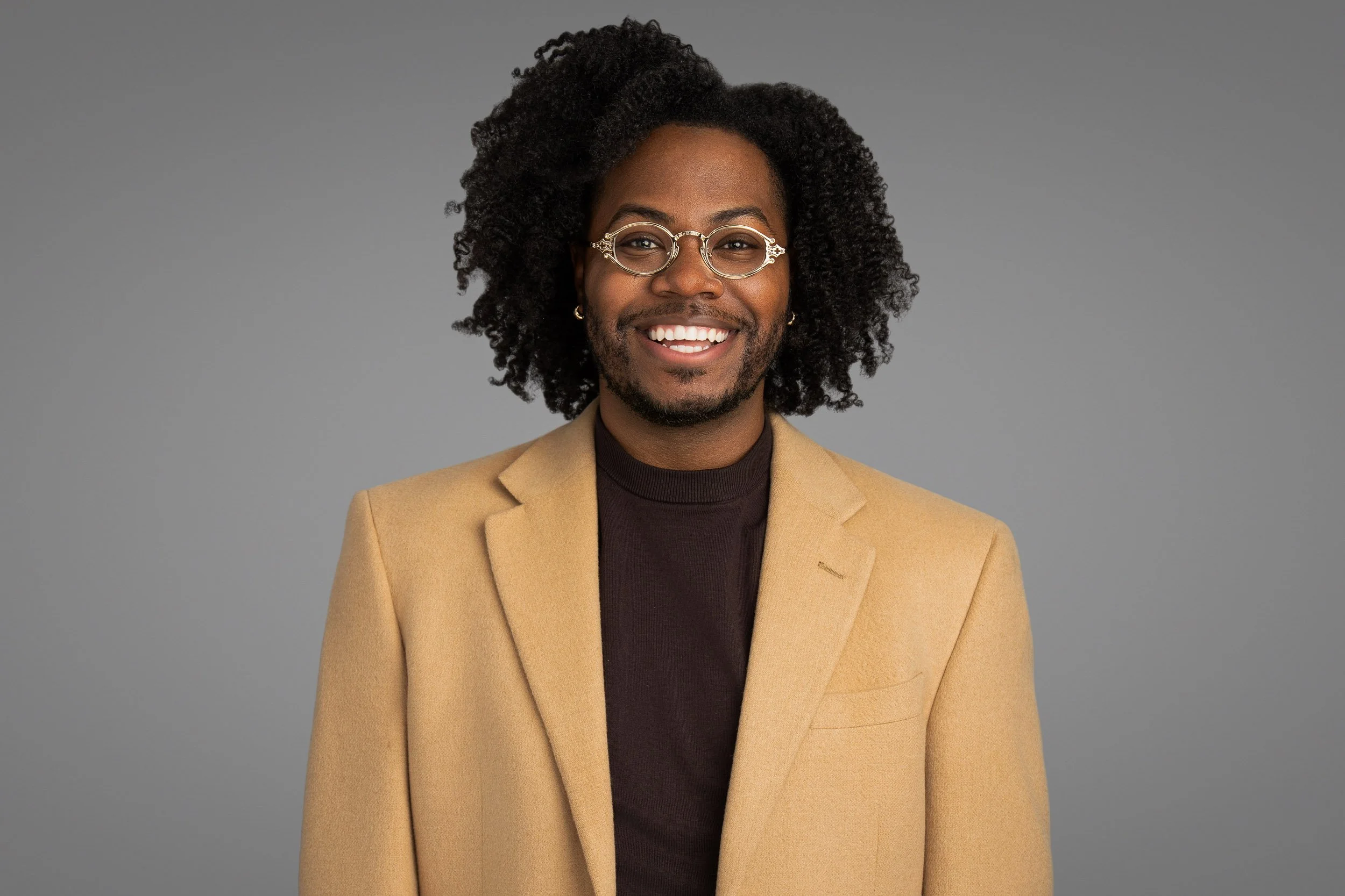 A smiling man with curly black hair, glasses, earrings, wearing a beige blazer over a dark shirt, against a gray background.