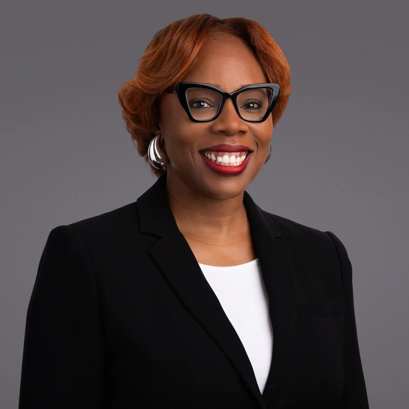 A smiling woman with short reddish hair, wearing glasses, large silver earrings, a black blazer, and a white shirt, against a gray background.
