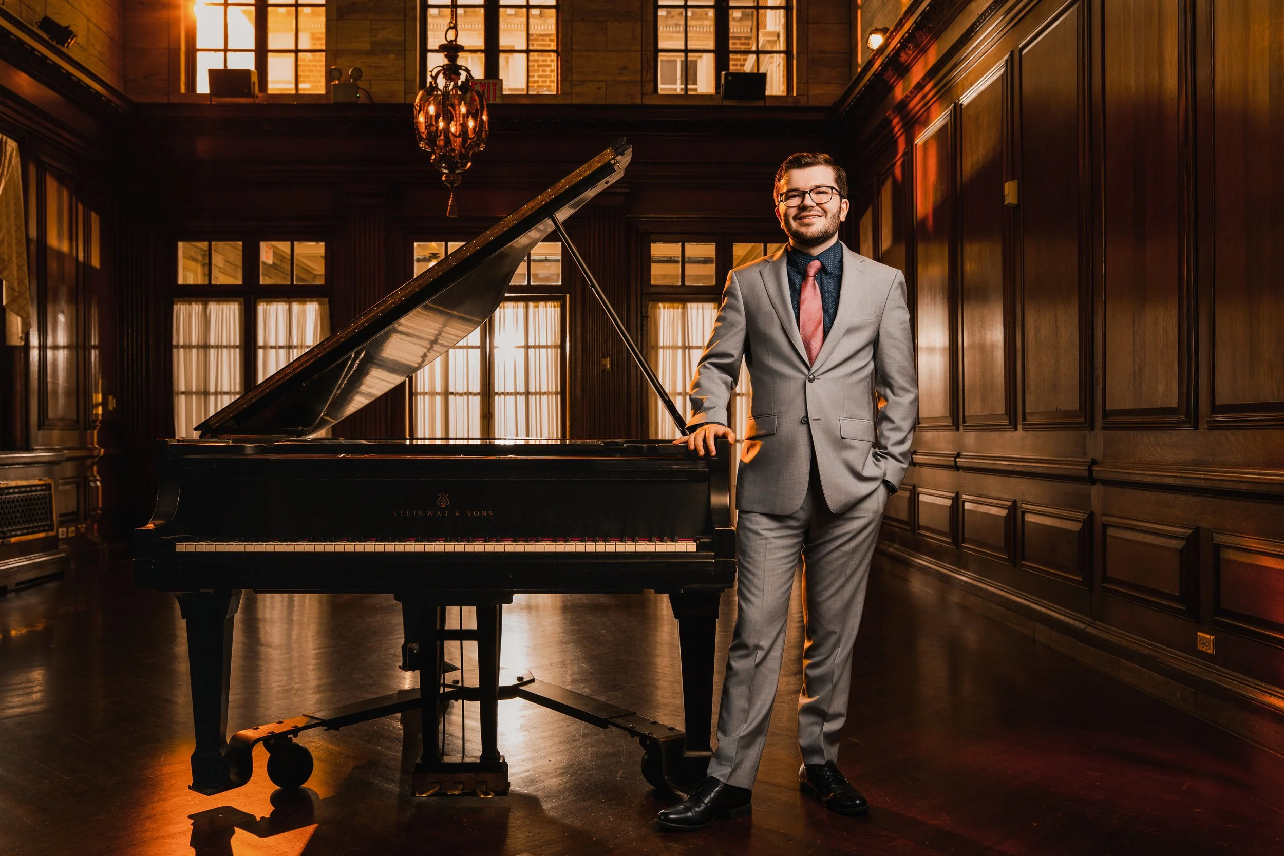 A man in a gray suit with glasses and a pink tie standing next to a black grand piano inside a wooden-paneled room with large windows and warm lighting.